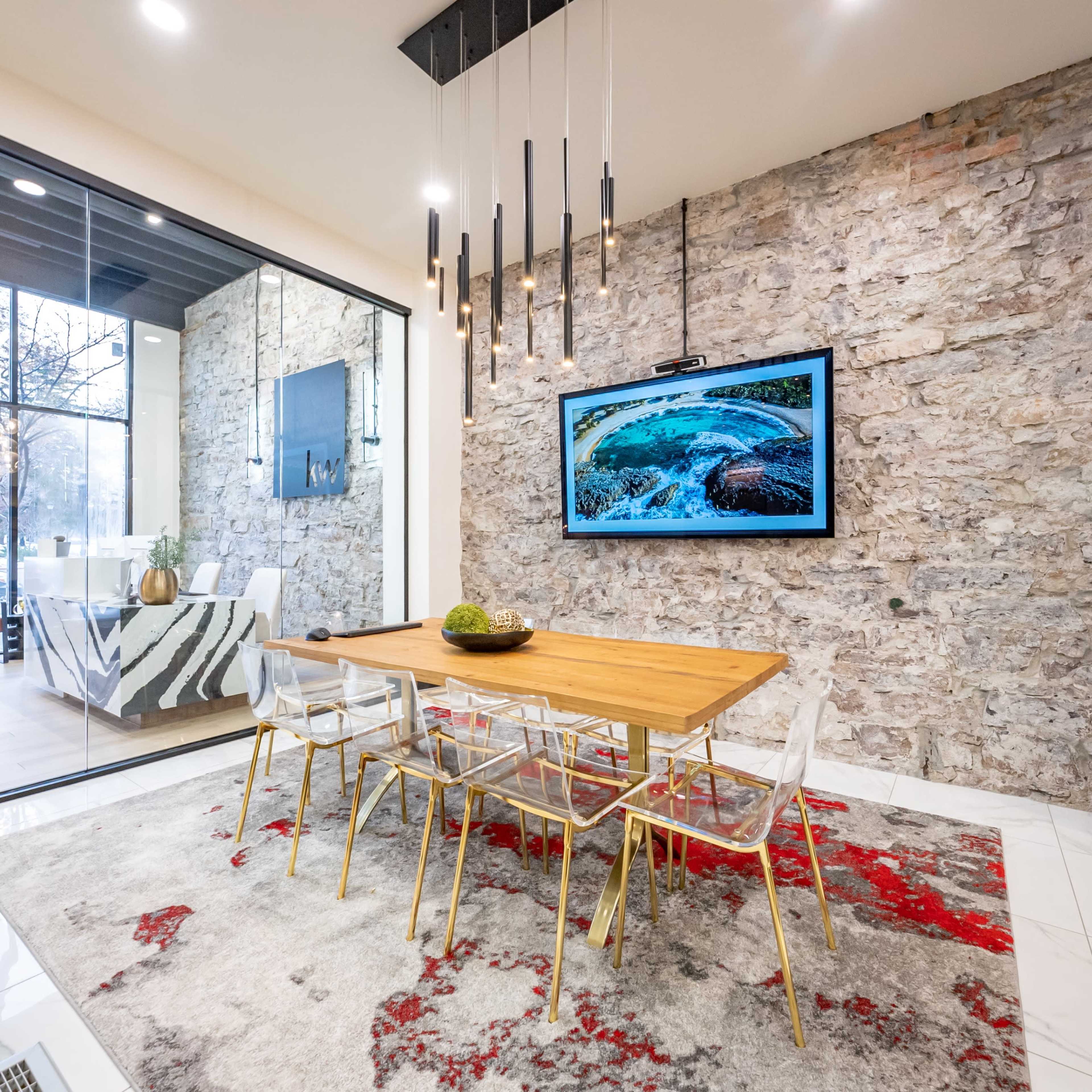 A modern dining area featuring a wooden table surrounded by clear acrylic chairs, set against a stone accent wall with a large television displaying an image.