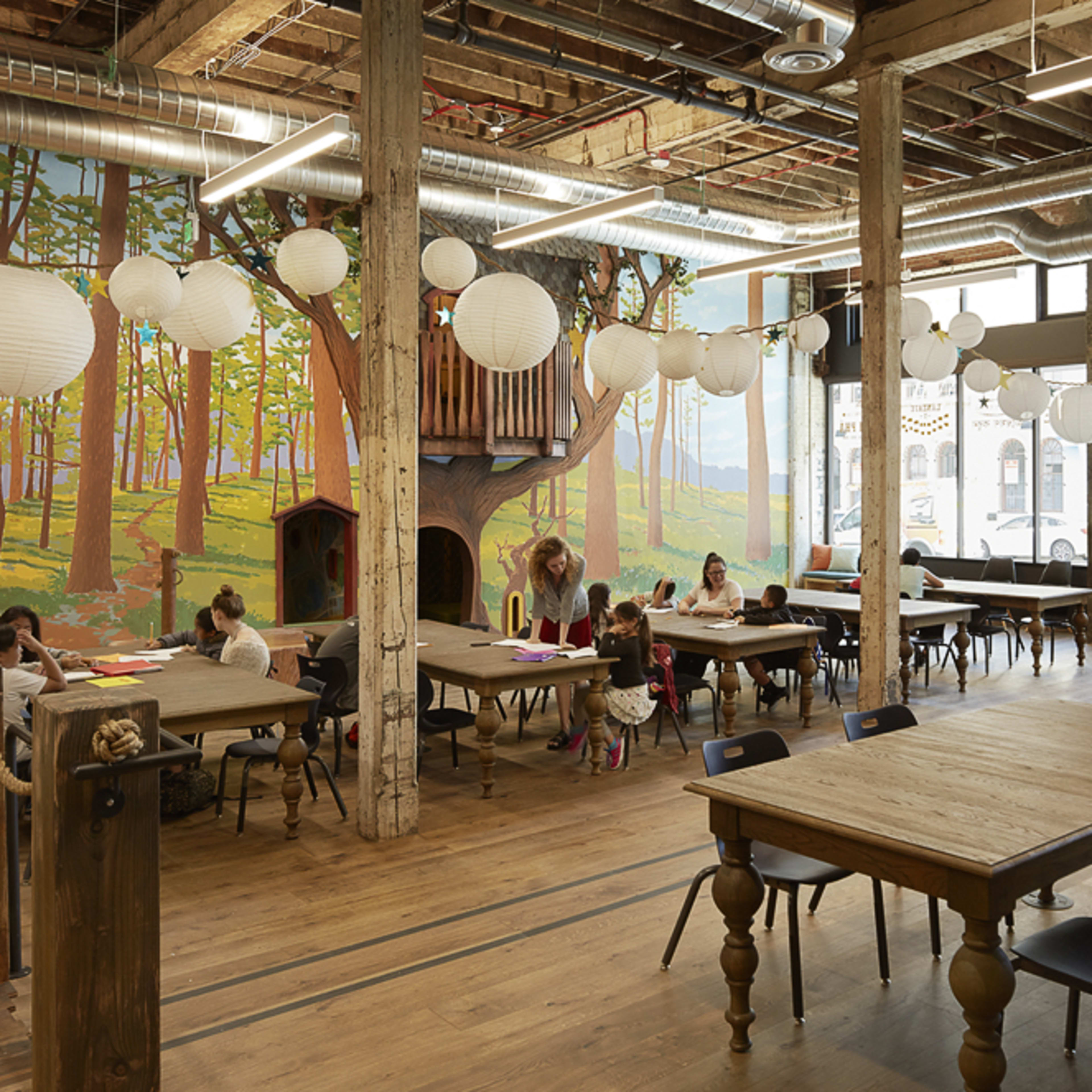 A spacious, well-lit interior of a creative workspace with large tables, wooden flooring, and a mural of trees on the far wall.