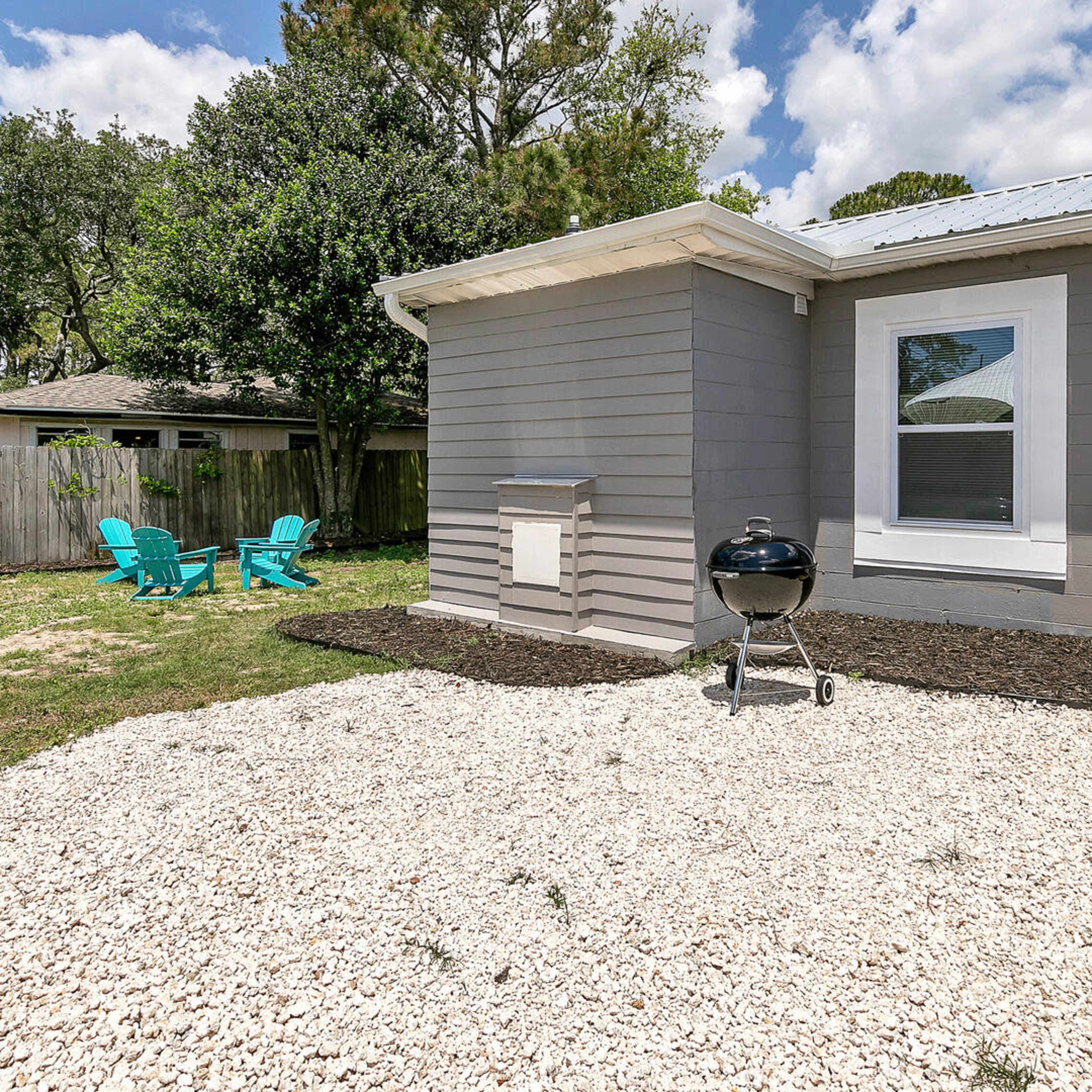 The side of a house featuring a gravel patio, a small grill, and two turquoise chairs in a grassy yard.