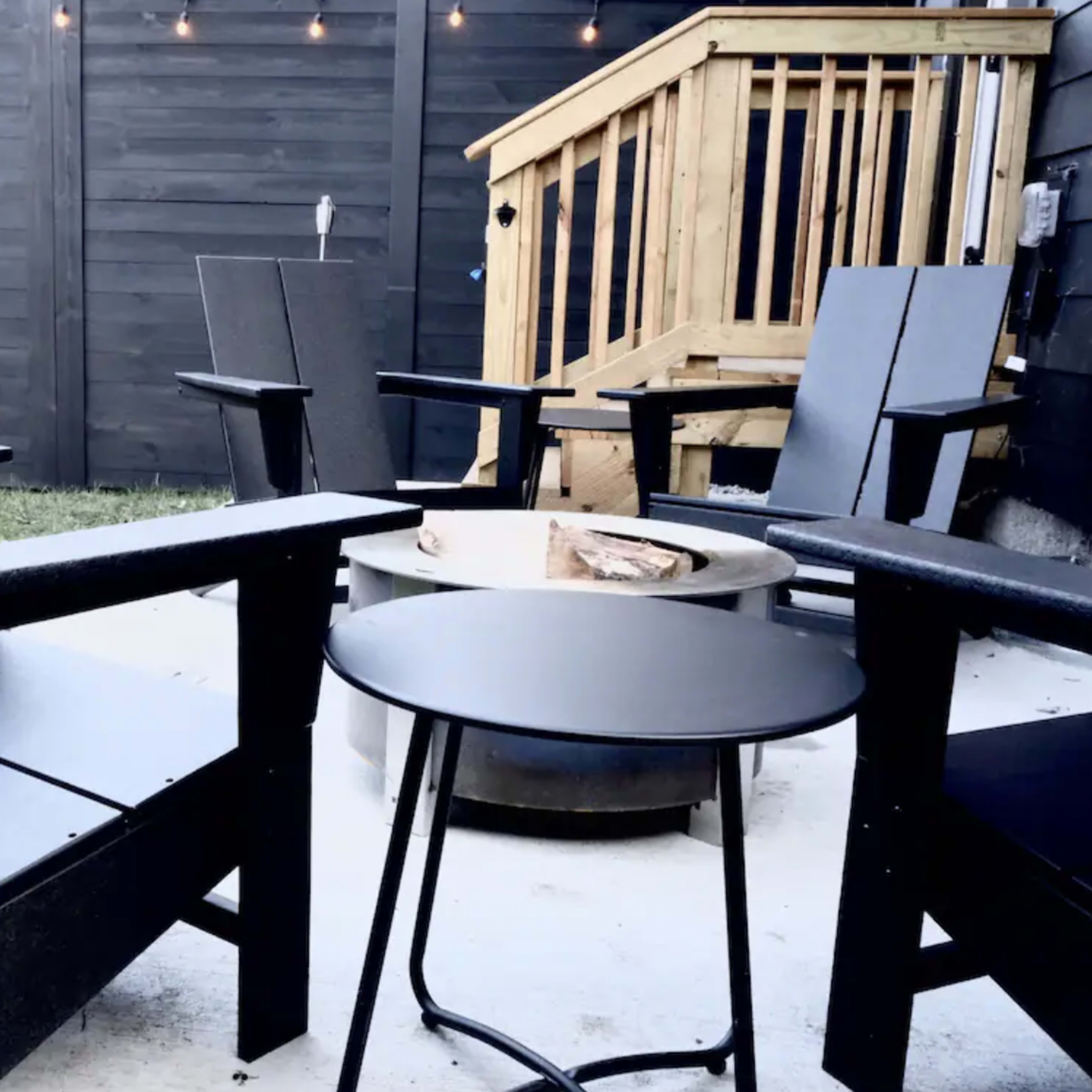 A patio area with four black Adirondack chairs arranged around a circular fire pit, along with a small black side table and wooden steps leading to a raised deck.