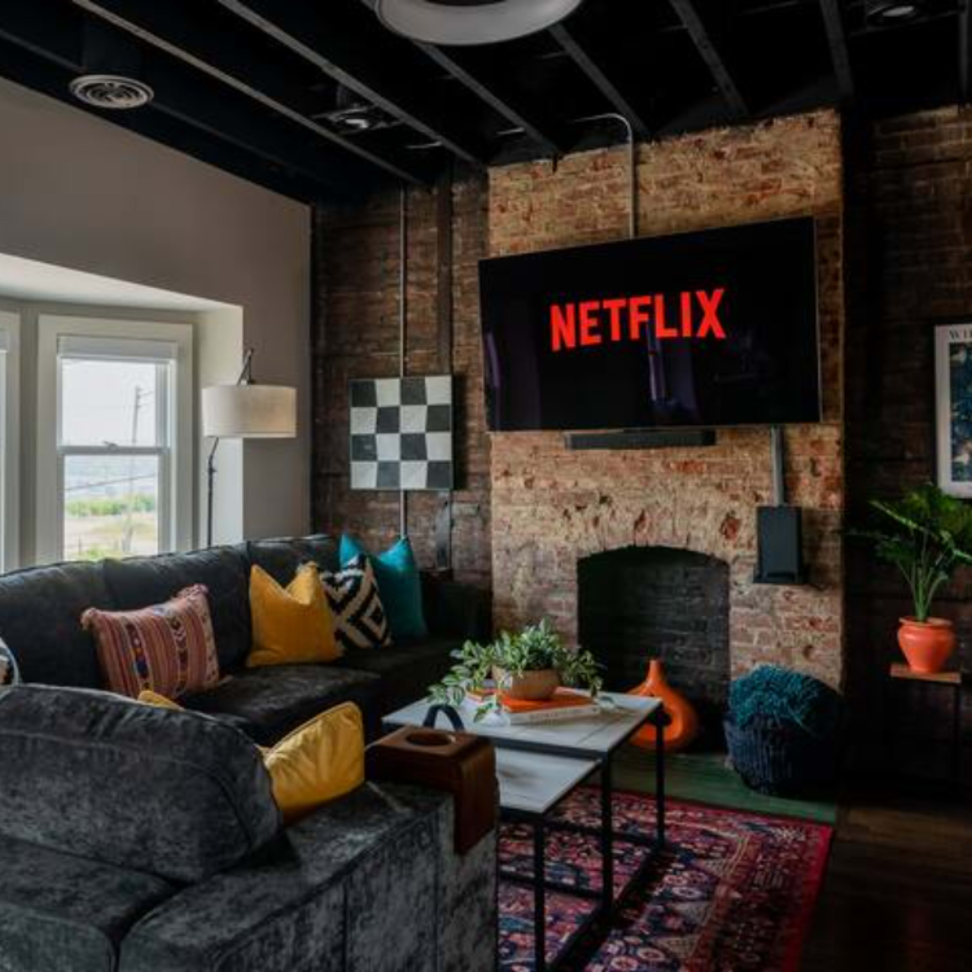 A modern living room features a large sectional sofa, a brick wall with a mounted television displaying the Netflix logo, and a bright window showcasing an outdoor view.