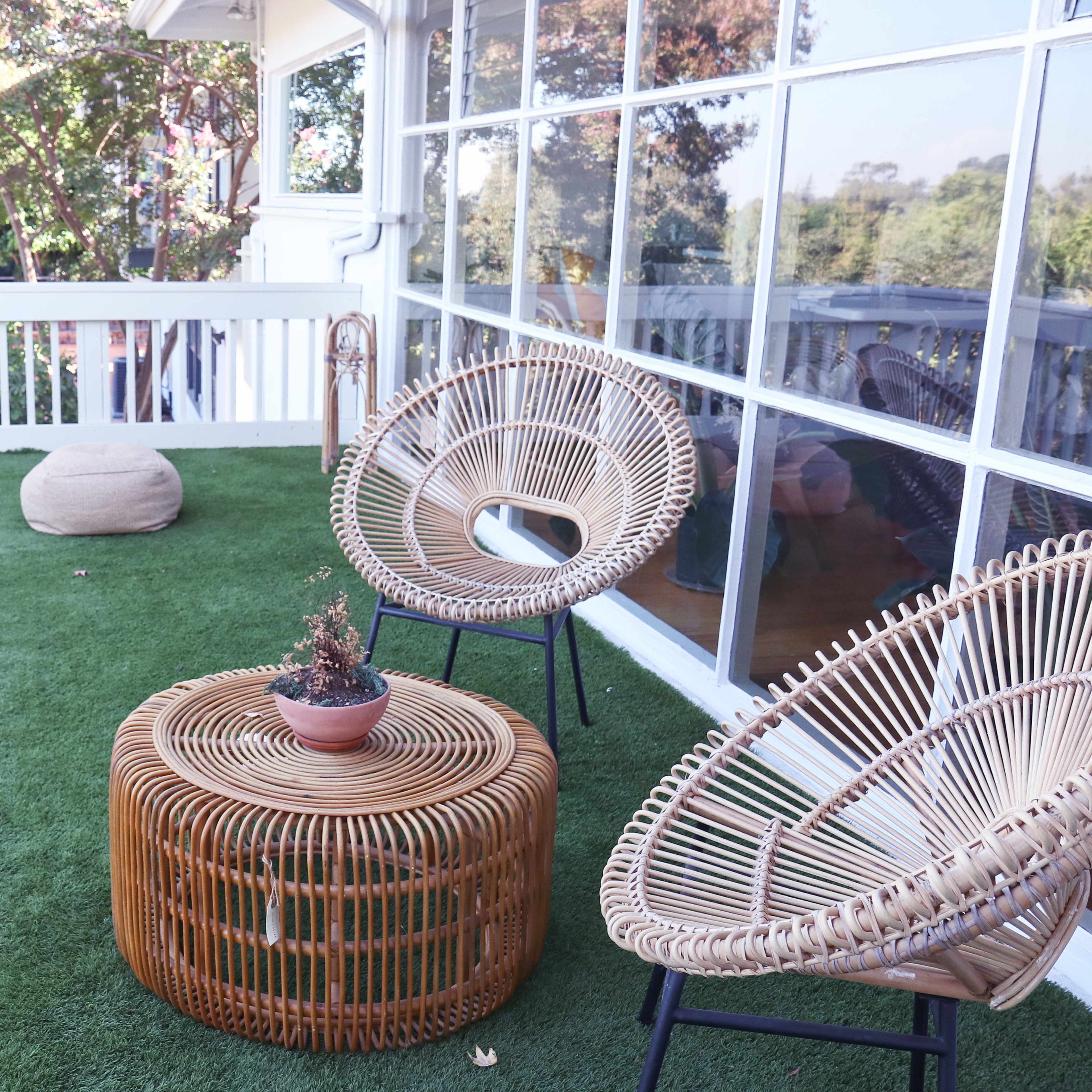A balcony features two woven chairs and a round wicker table on artificial grass, surrounded by greenery.