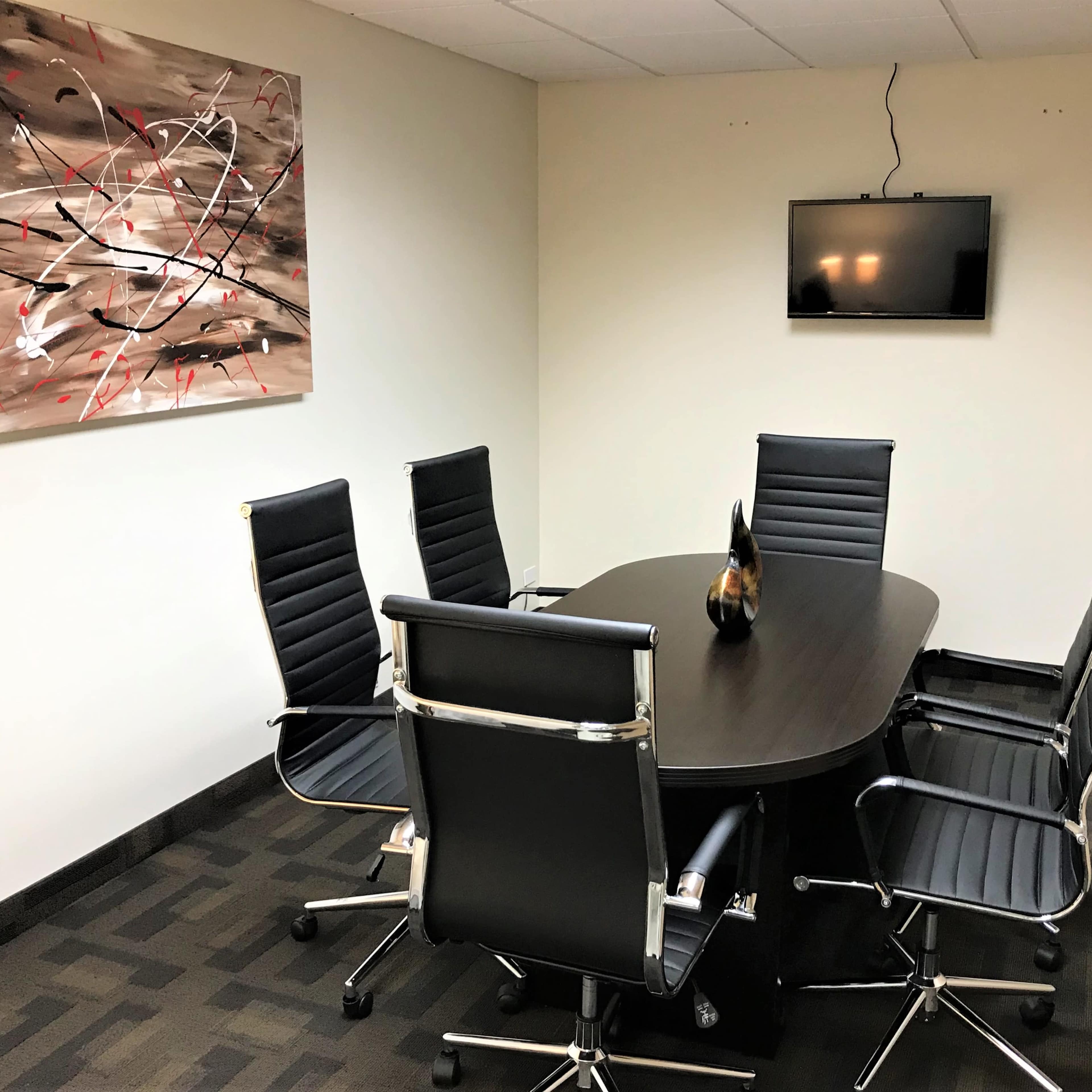 A conference room features a large oval table surrounded by six black leather chairs, with an abstract painting on the wall and a television mounted above.