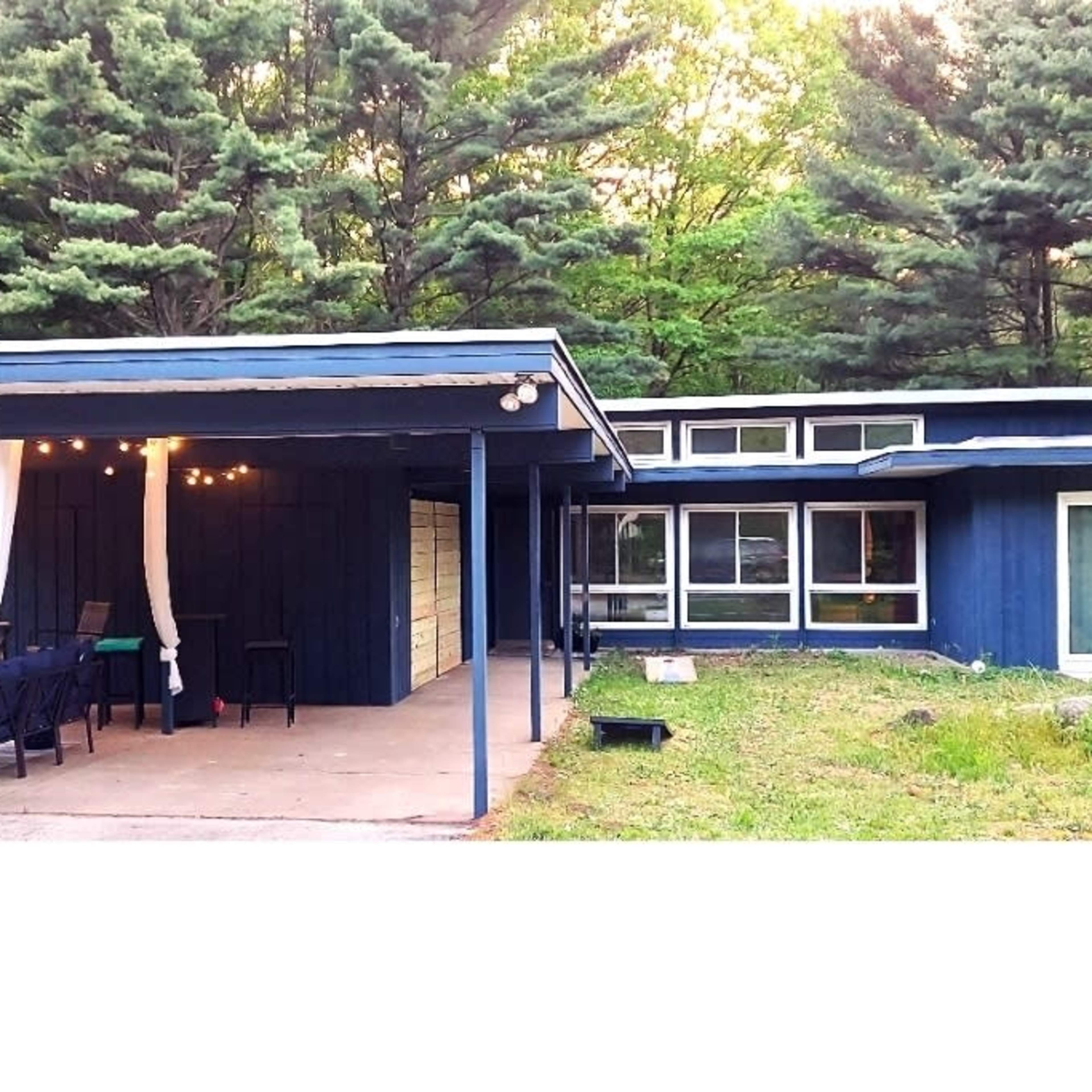 A blue and white cabin features a covered patio with string lights, surrounded by tall trees.