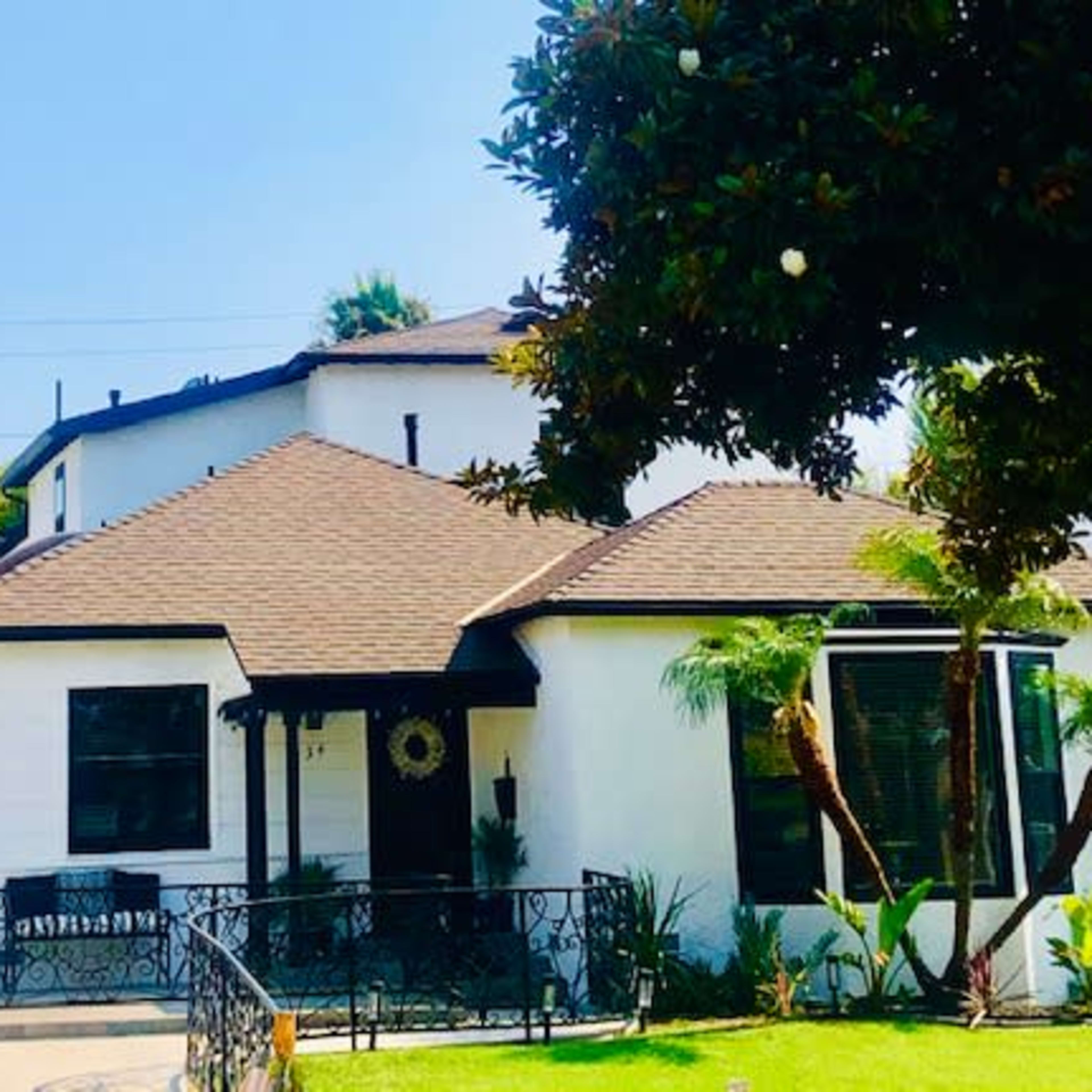 A white house with a brown, multi-angled roof, surrounded by greenery and featuring a wrought iron gate.