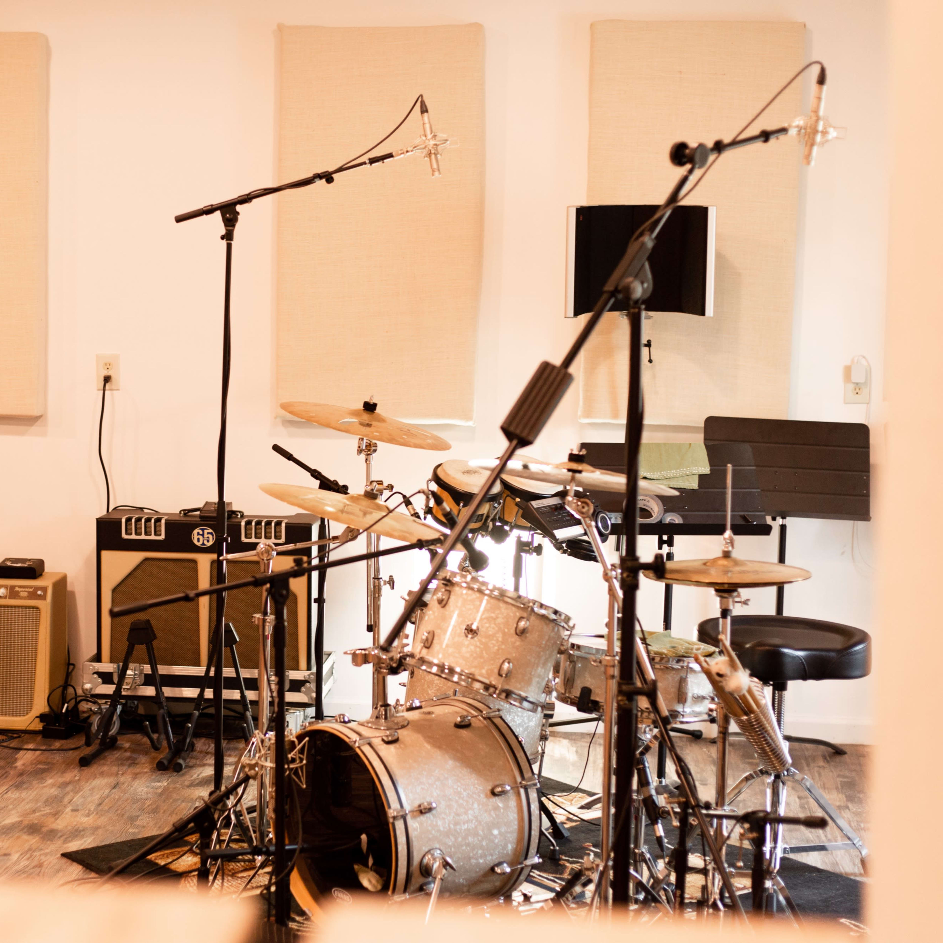 A music studio is equipped with microphones, amplifiers, and a drum set in front of soundproof panels.