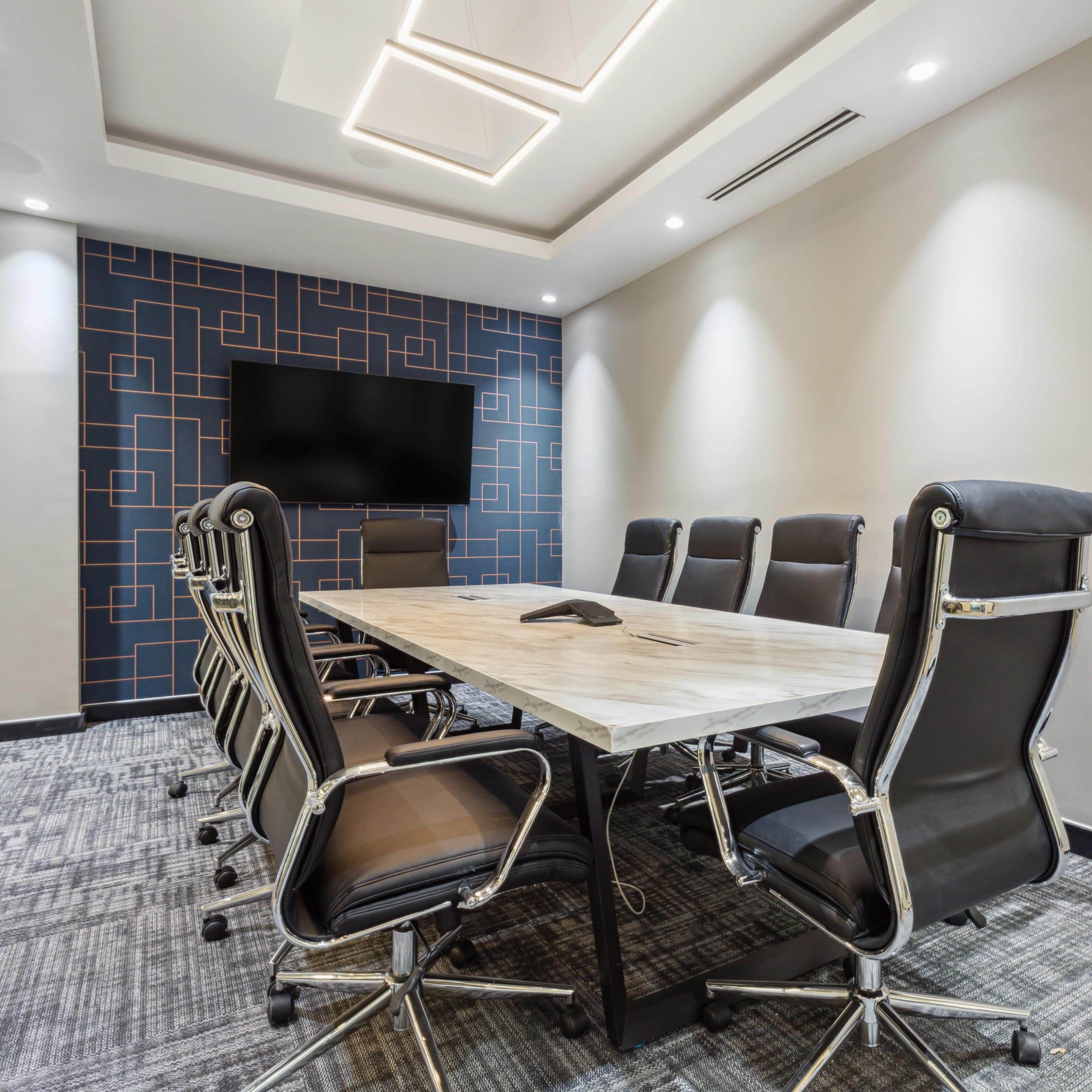 A modern conference room features a rectangular table surrounded by ergonomic chairs and a wall-mounted display.
