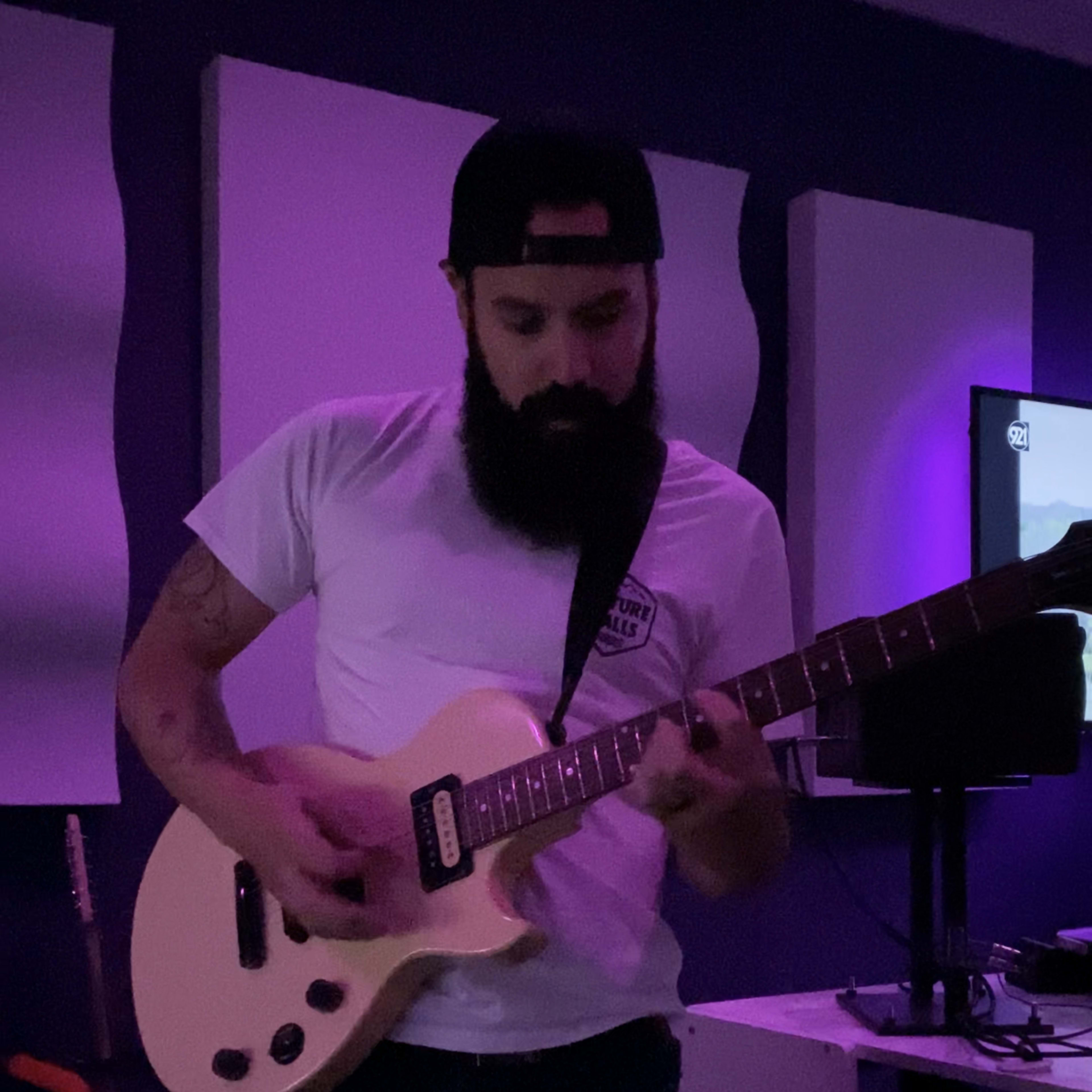 A man with a beard plays an electric guitar in a dimly lit room with purple lighting.