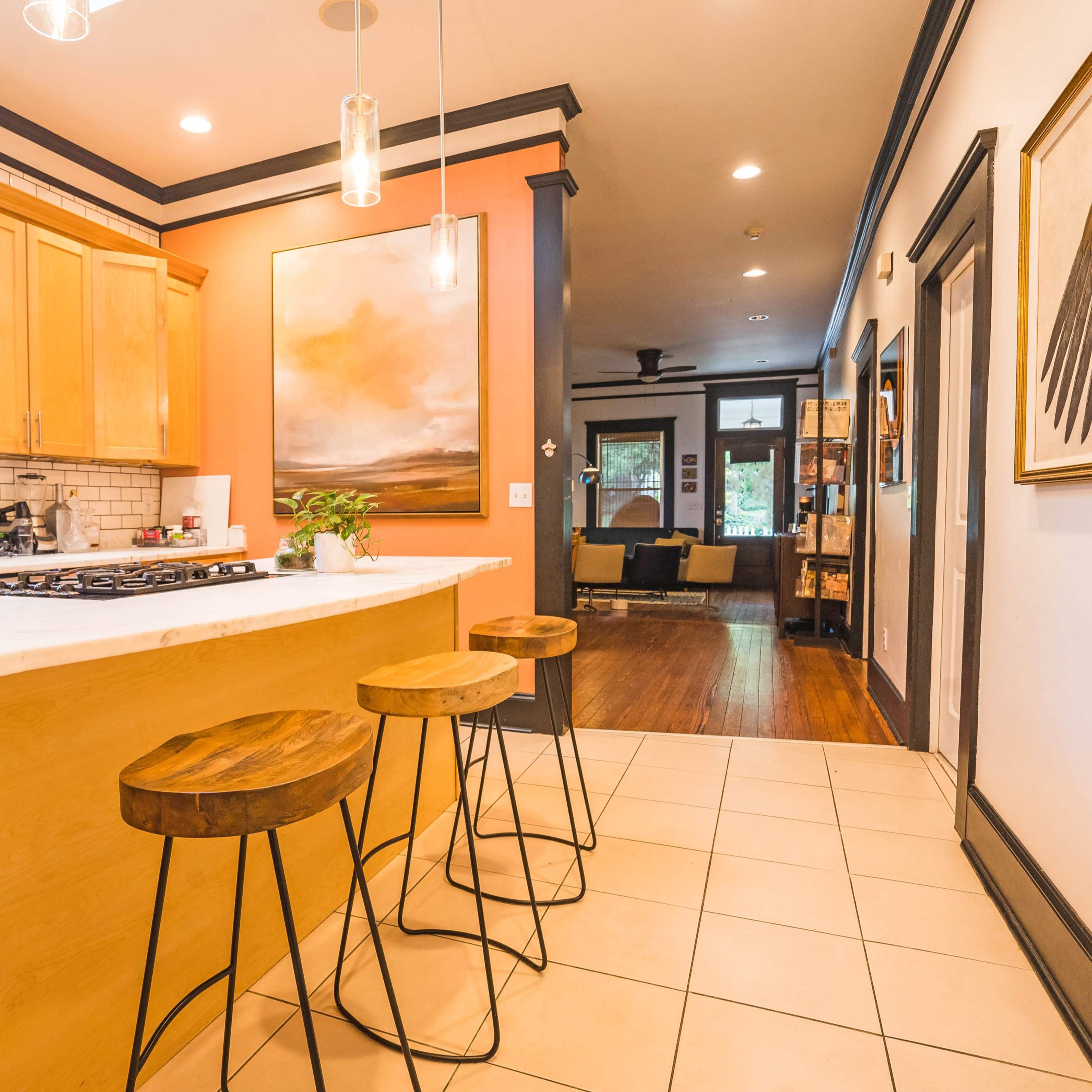A contemporary kitchen with wooden cabinetry, a marble countertop, and three metal bar stools, leading into a living area with a sofa and artwork on the walls.