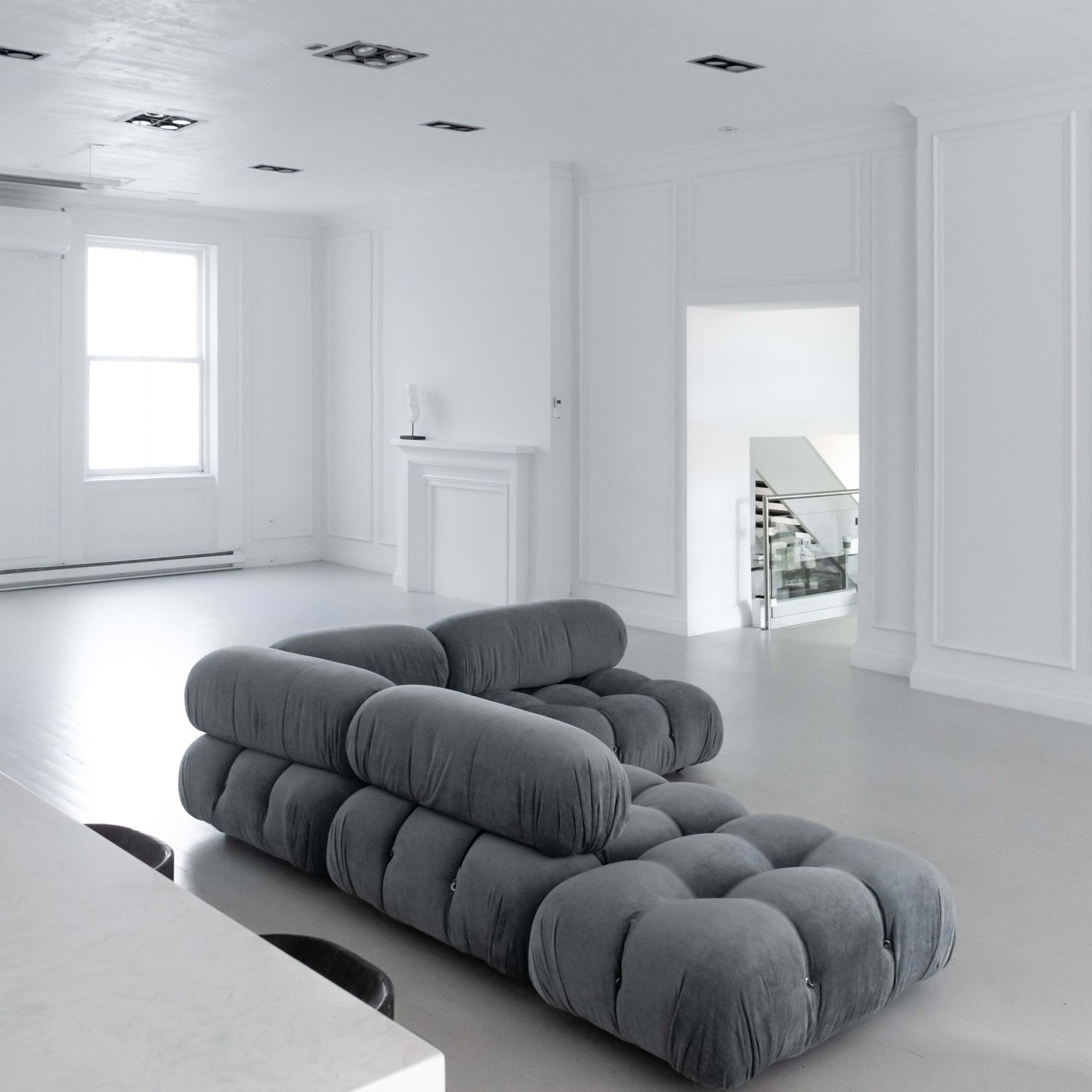 A gray tufted sofa is positioned in a spacious, mostly white room with large windows and minimal furniture.
