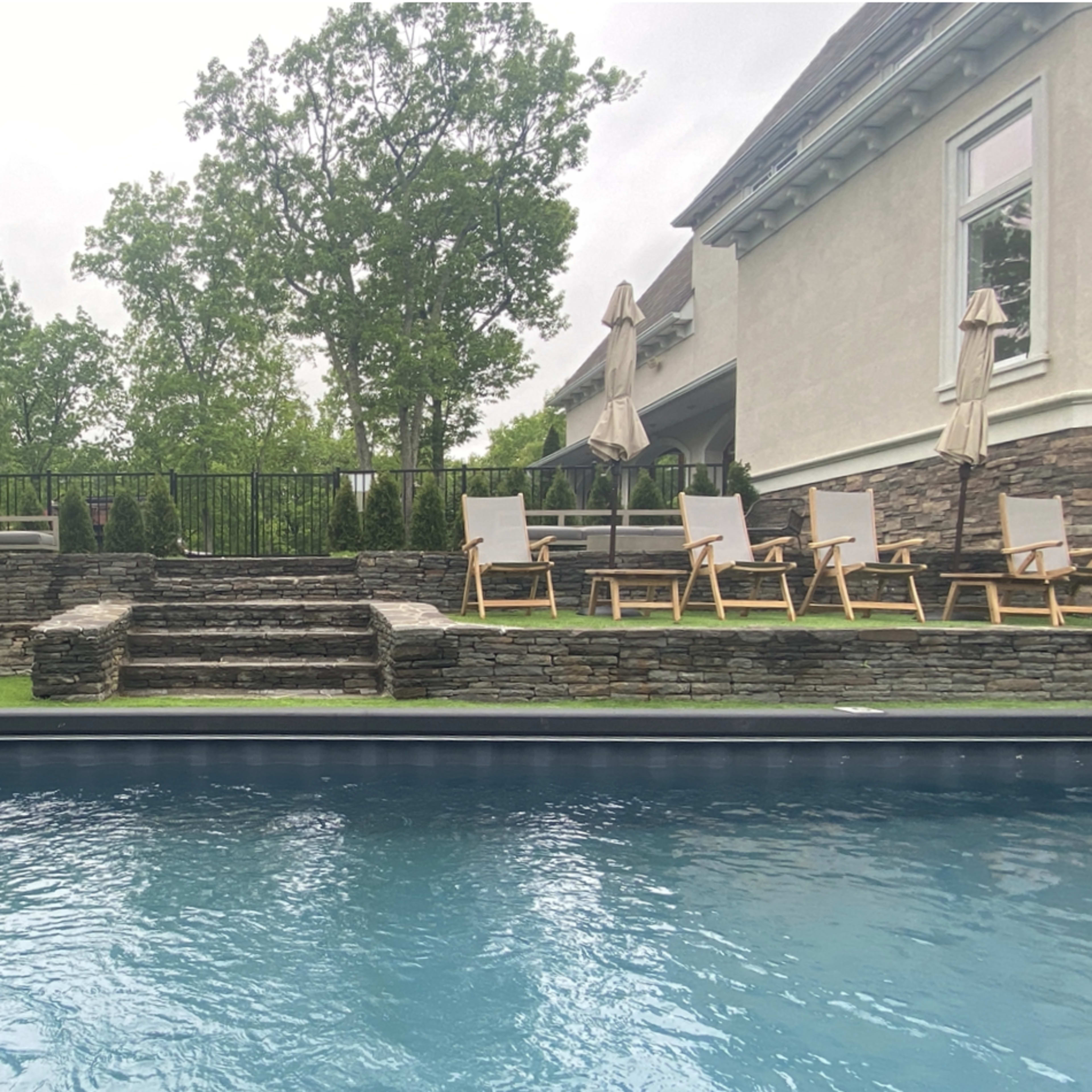 A swimming pool with wooden lounge chairs and umbrellas arranged beside a stone terrace with steps leading up to a grassy area.