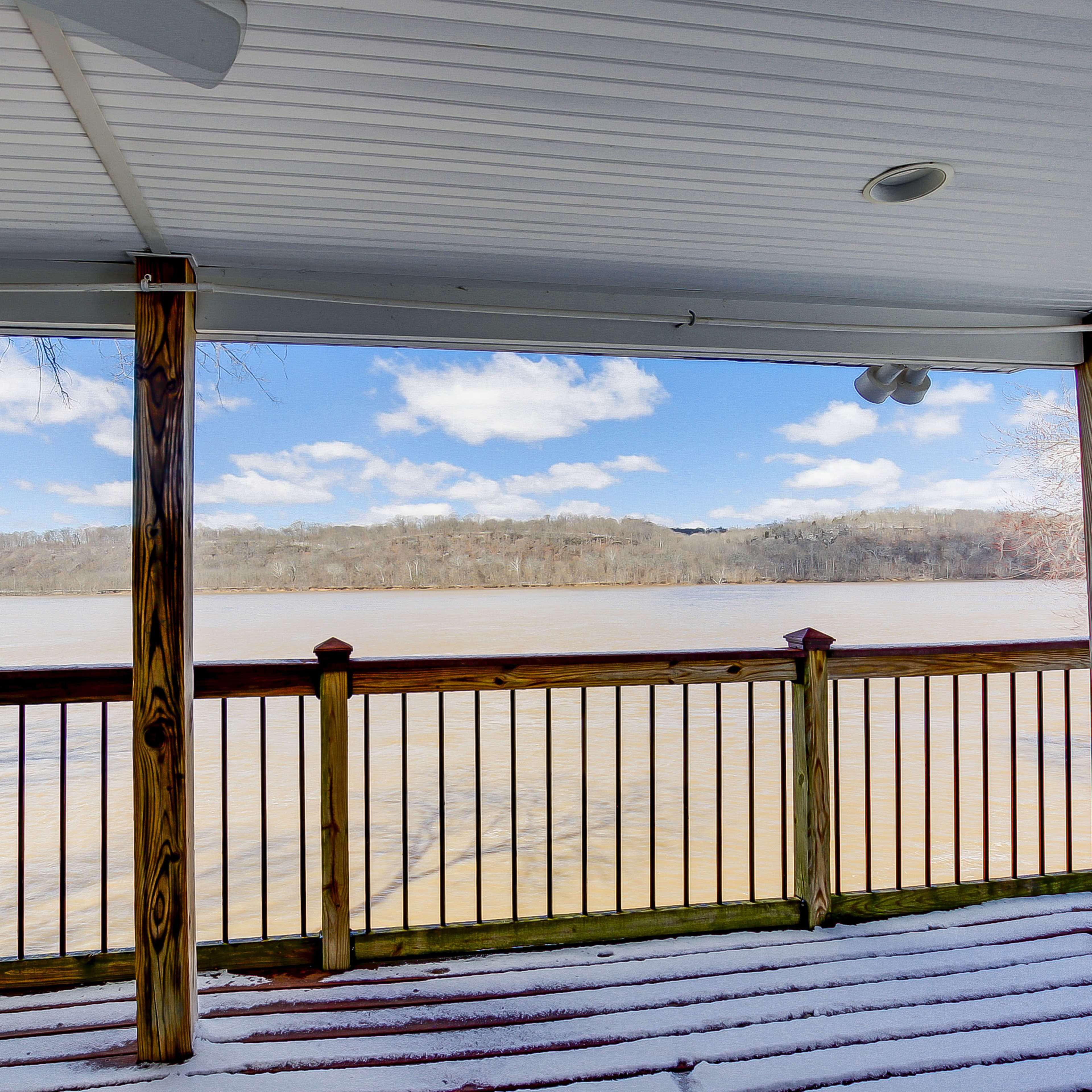 A covered porch with wooden railings overlooks a river bordered by trees under a cloudy sky.