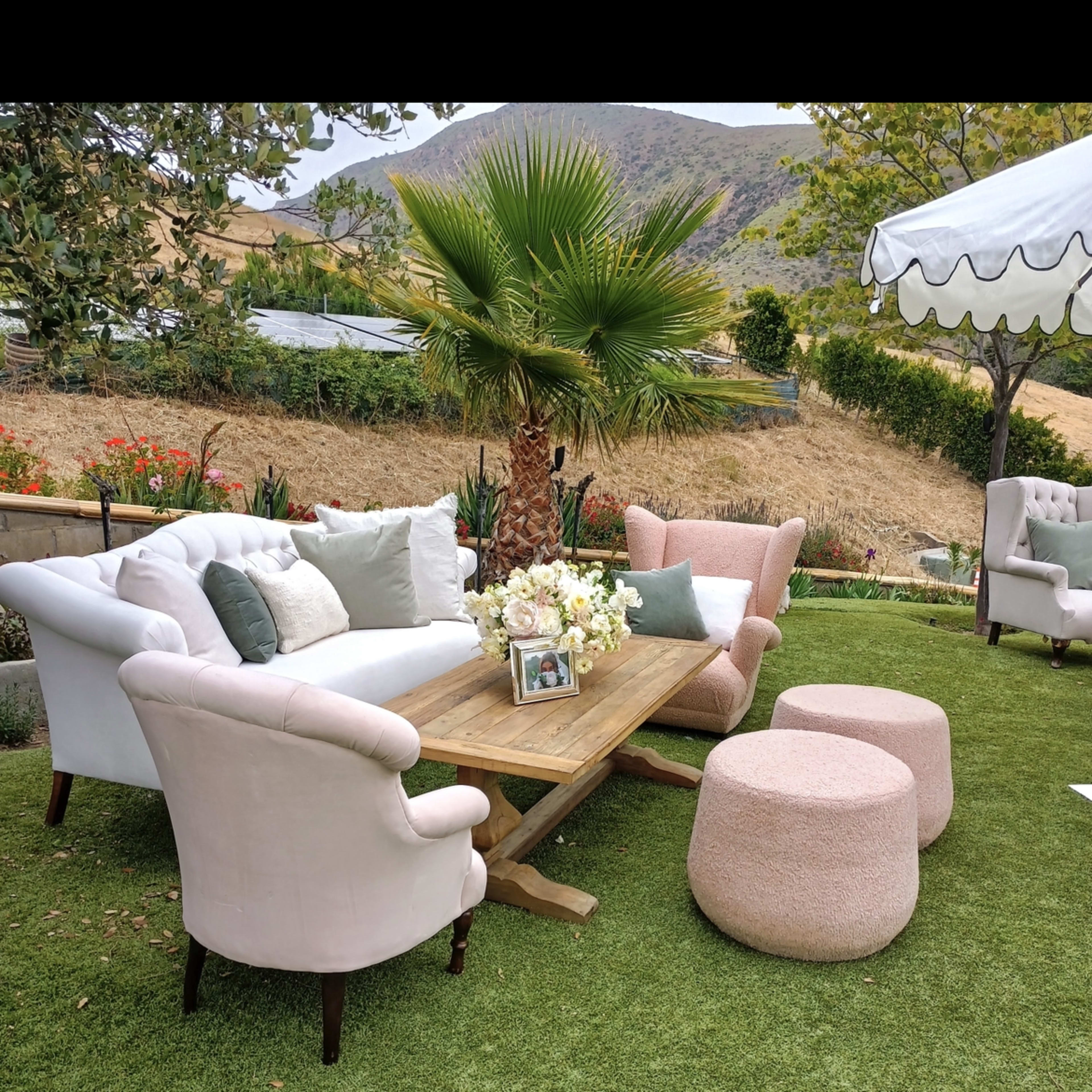 A set of upholstered outdoor furniture, including a wooden table, is arranged on green grass with a palm tree and decorative plants in the background.