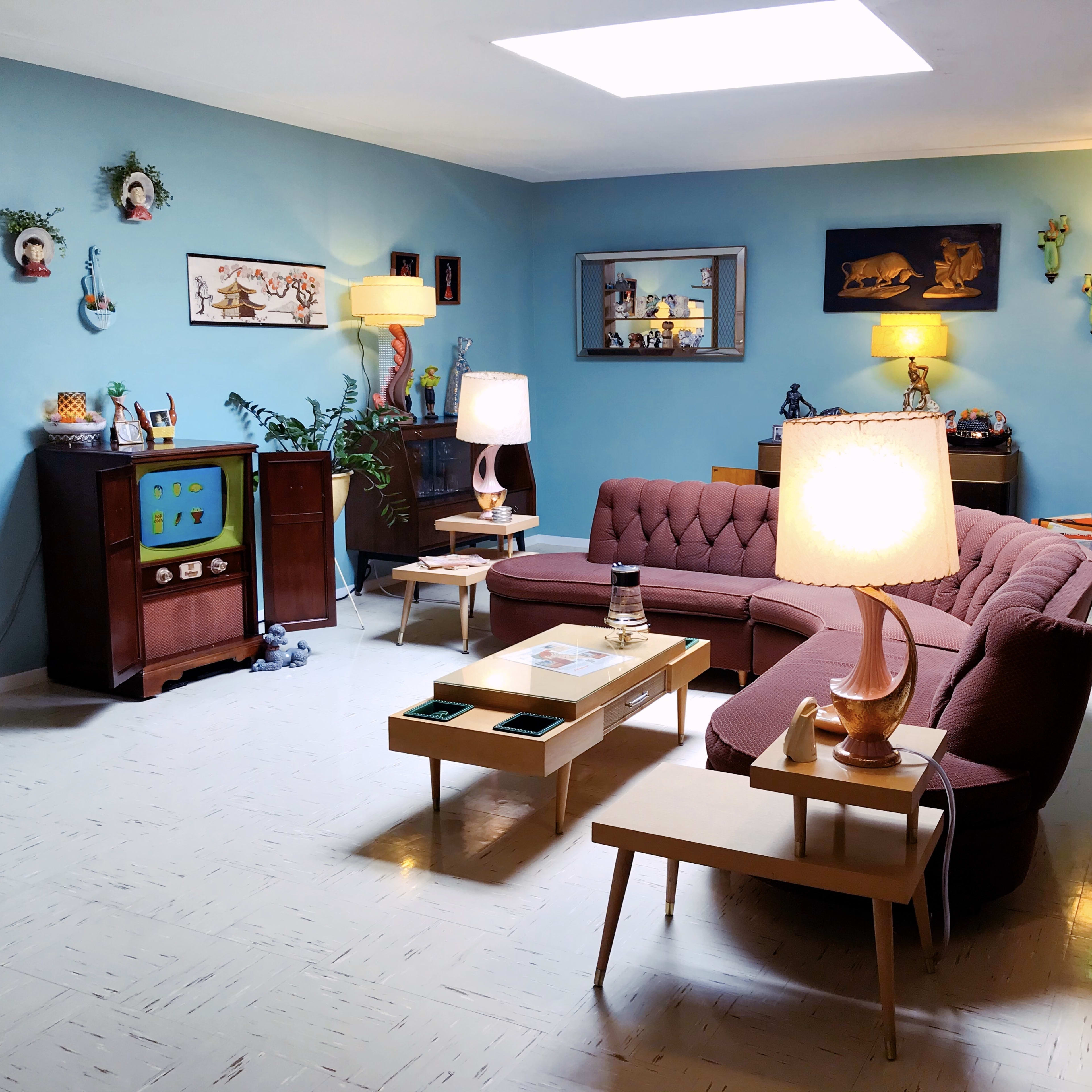 A brightly lit retro living room featuring a pink sectional sofa, a vintage television set, wooden side tables, and various decorative items on the walls.