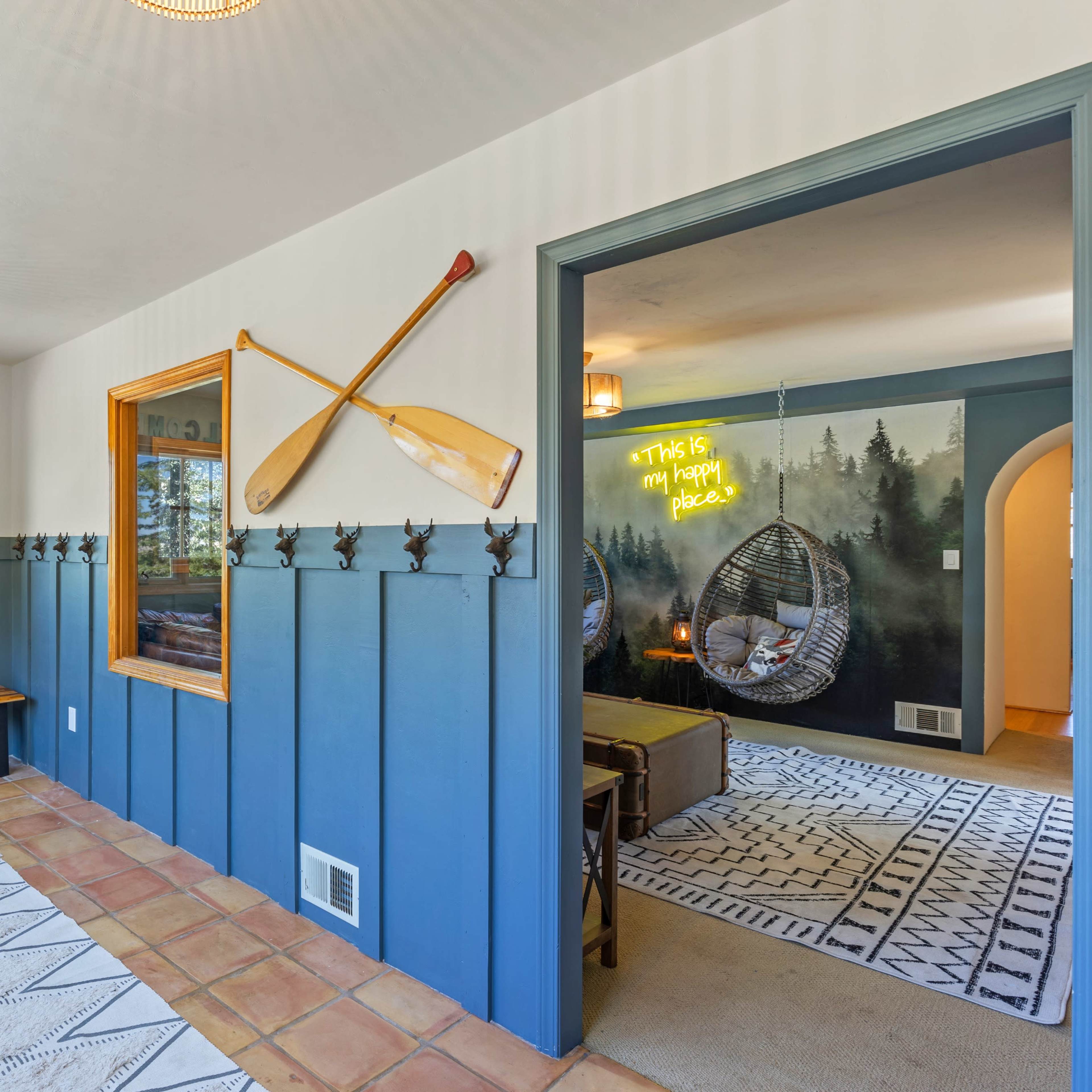 An entryway with a wooden bench, coat hooks on the wall, and a bright room visible through an open doorway featuring hanging chairs and decorative wall art.