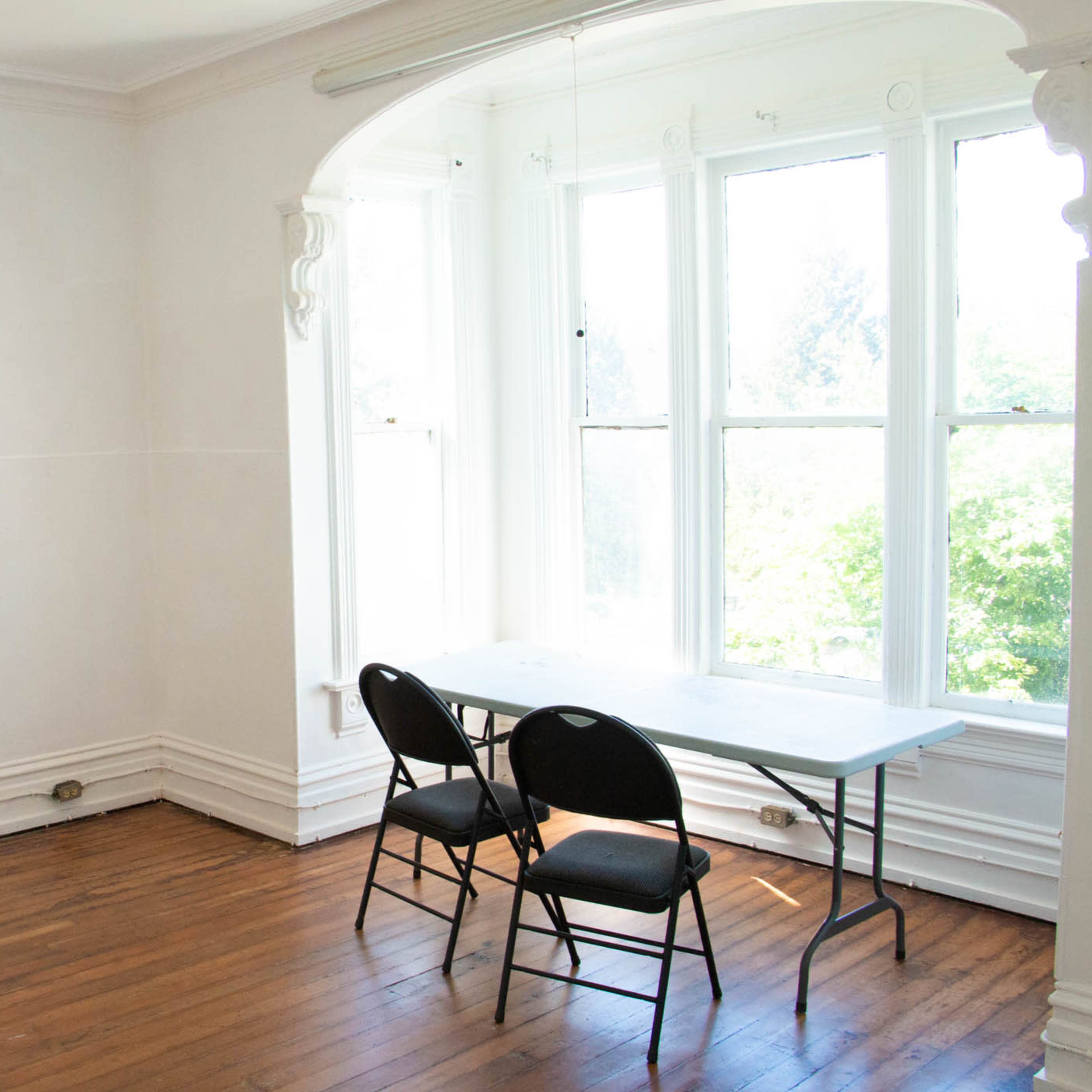 A sparsely furnished room features a table and two black chairs near a large window with a view of greenery outside.