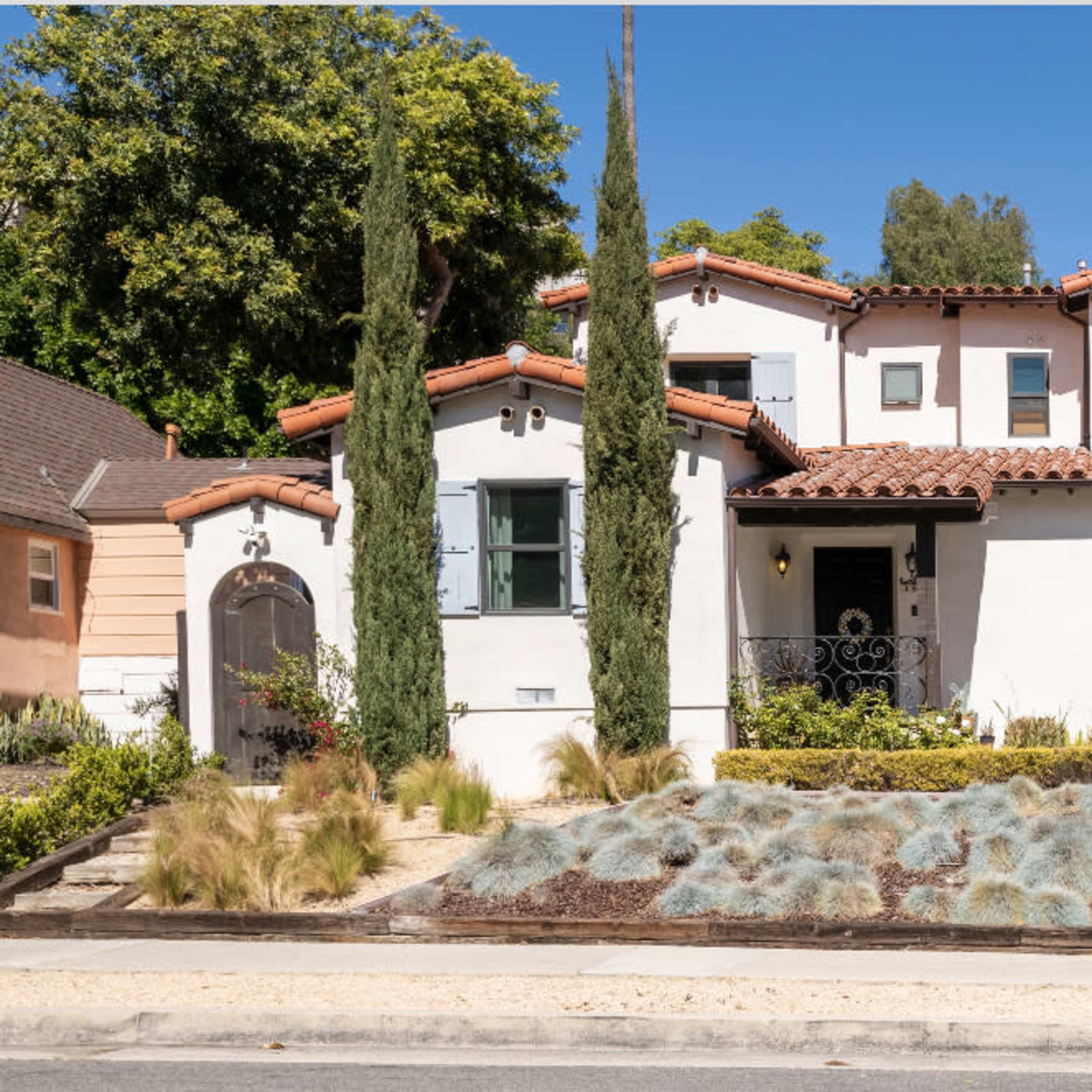 A two-story Mediterranean-style house with a terracotta roof, surrounded by neatly arranged landscaping and several tall cypress trees.