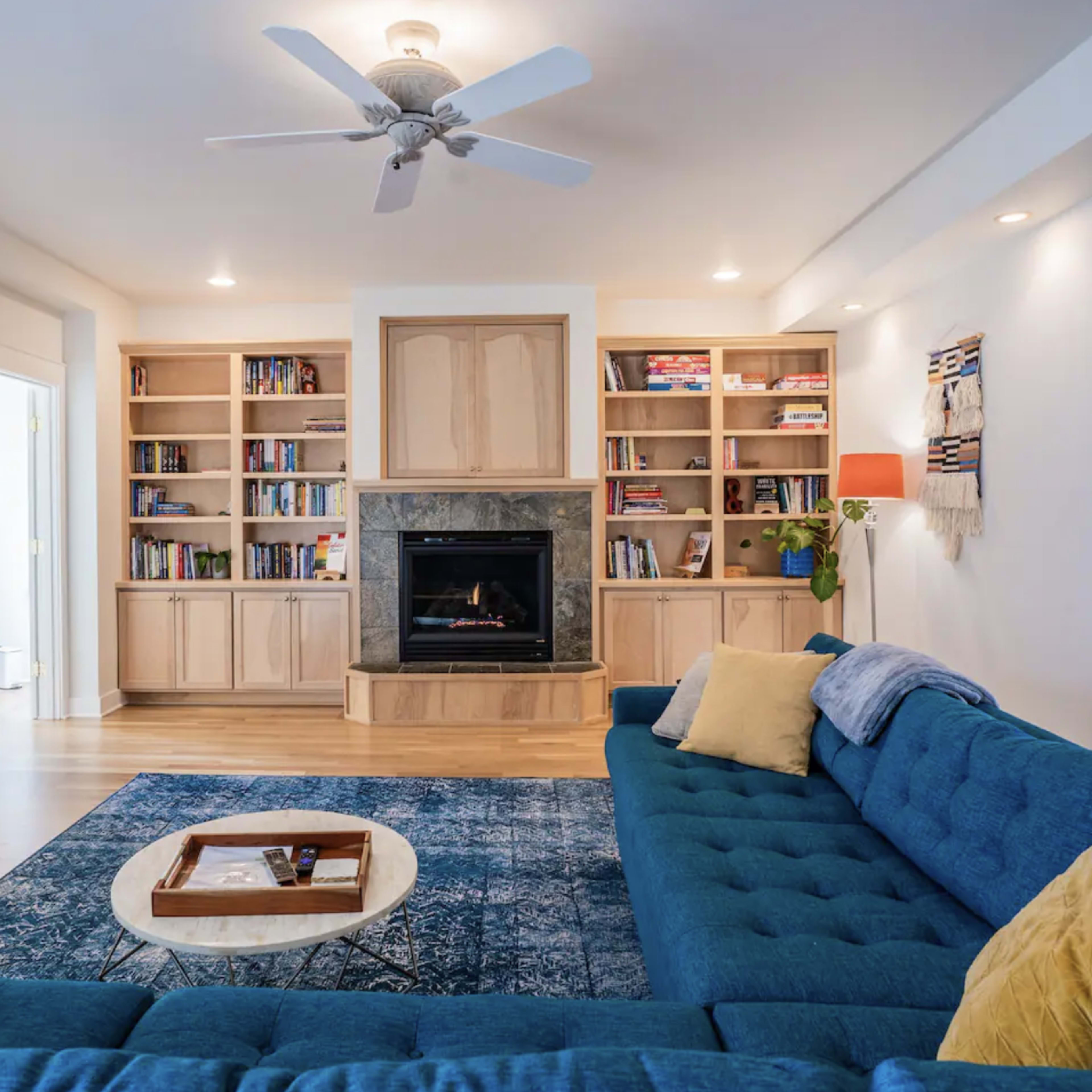 A living room features a blue sectional sofa, a fireplace surrounded by built-in bookshelves, and a decorative coffee table on a patterned rug.