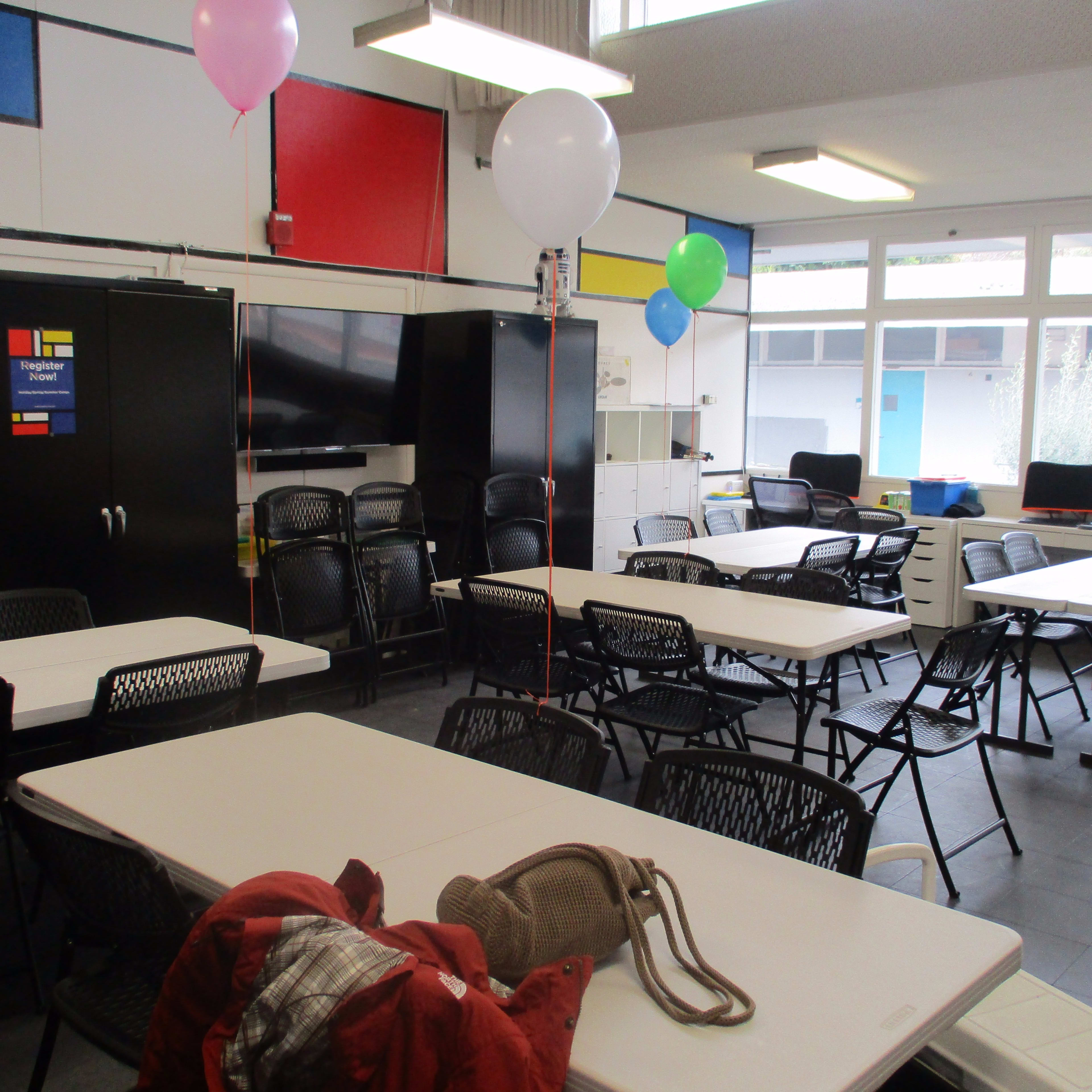 A room with several tables and chairs, decorated with balloons, and featuring a television and storage units.