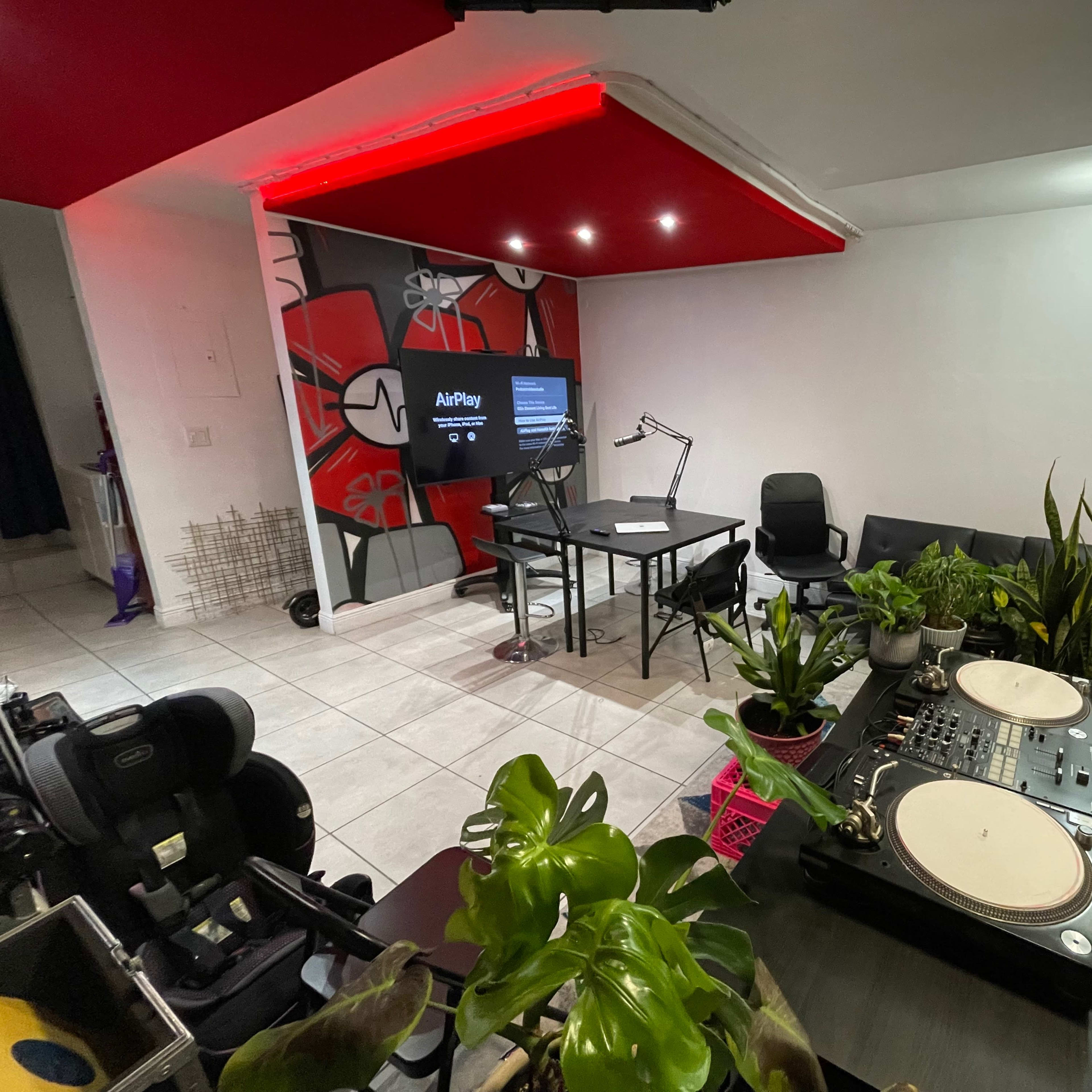 A stylish room set up with a DJ table, a large screen displaying "AirPlay," several plants, and contemporary seating arrangements.