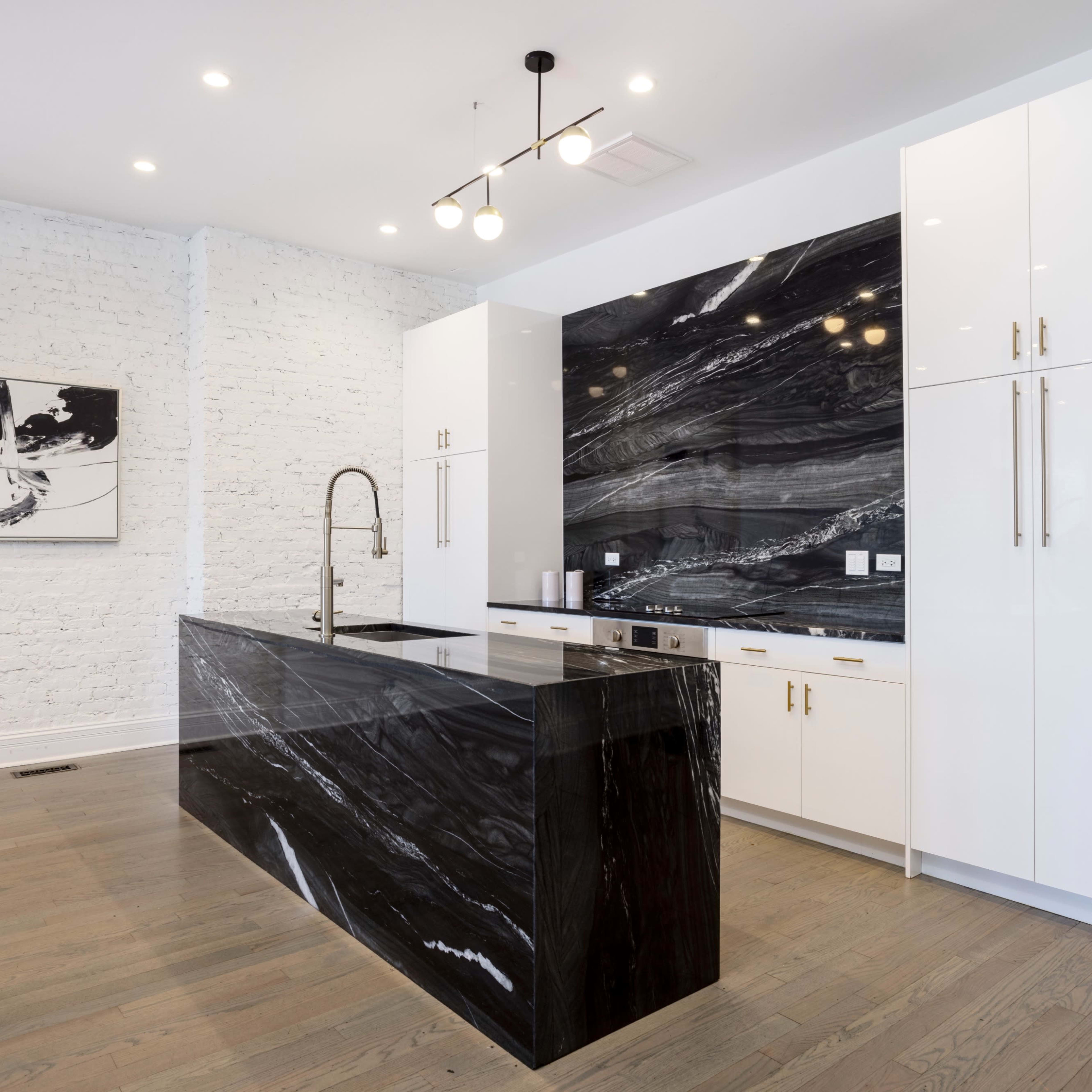 A modern kitchen featuring a black marble island with a sleek countertop and white cabinetry against a textured white brick wall.