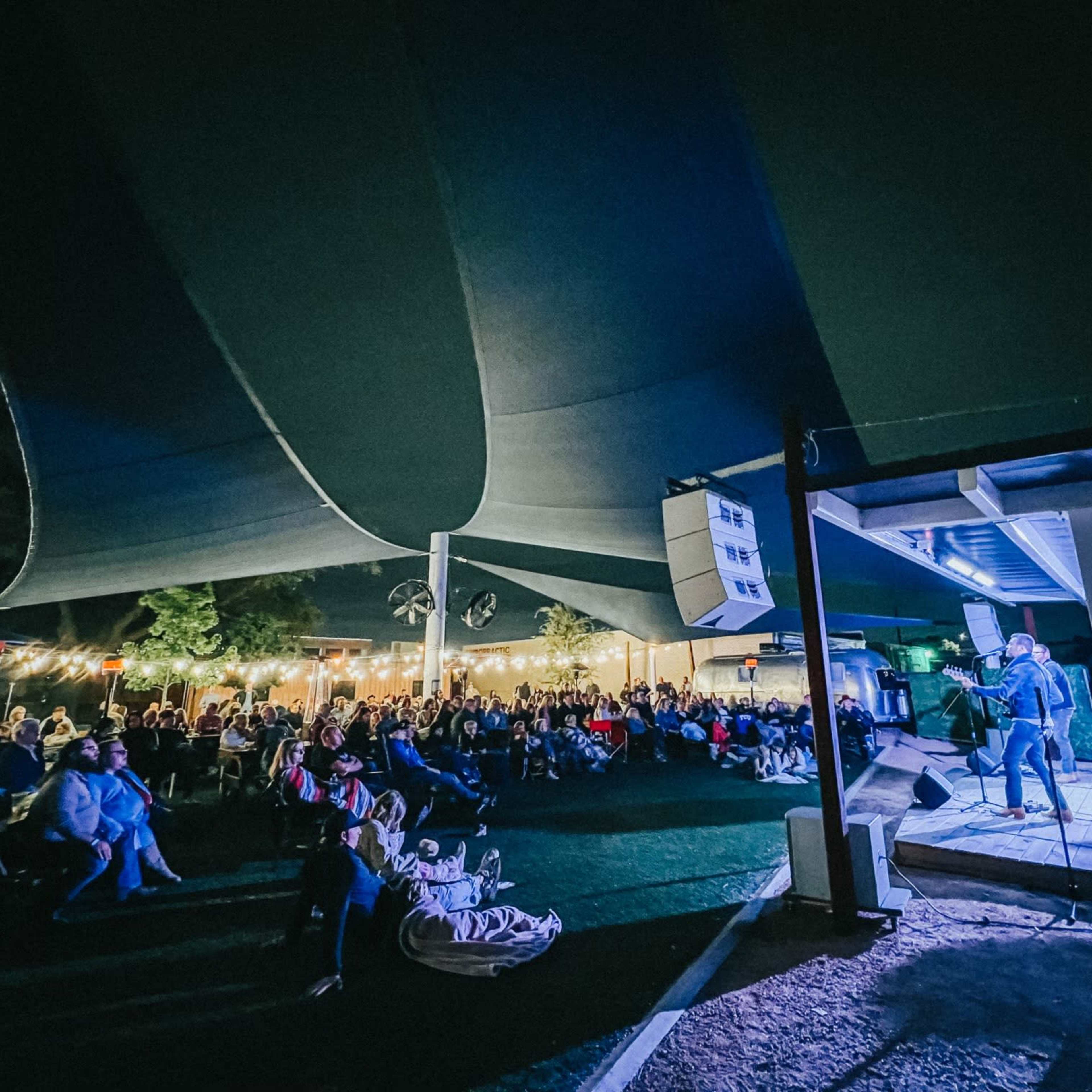 A musician performs on stage in front of a large audience under a covered outdoor venue at night.