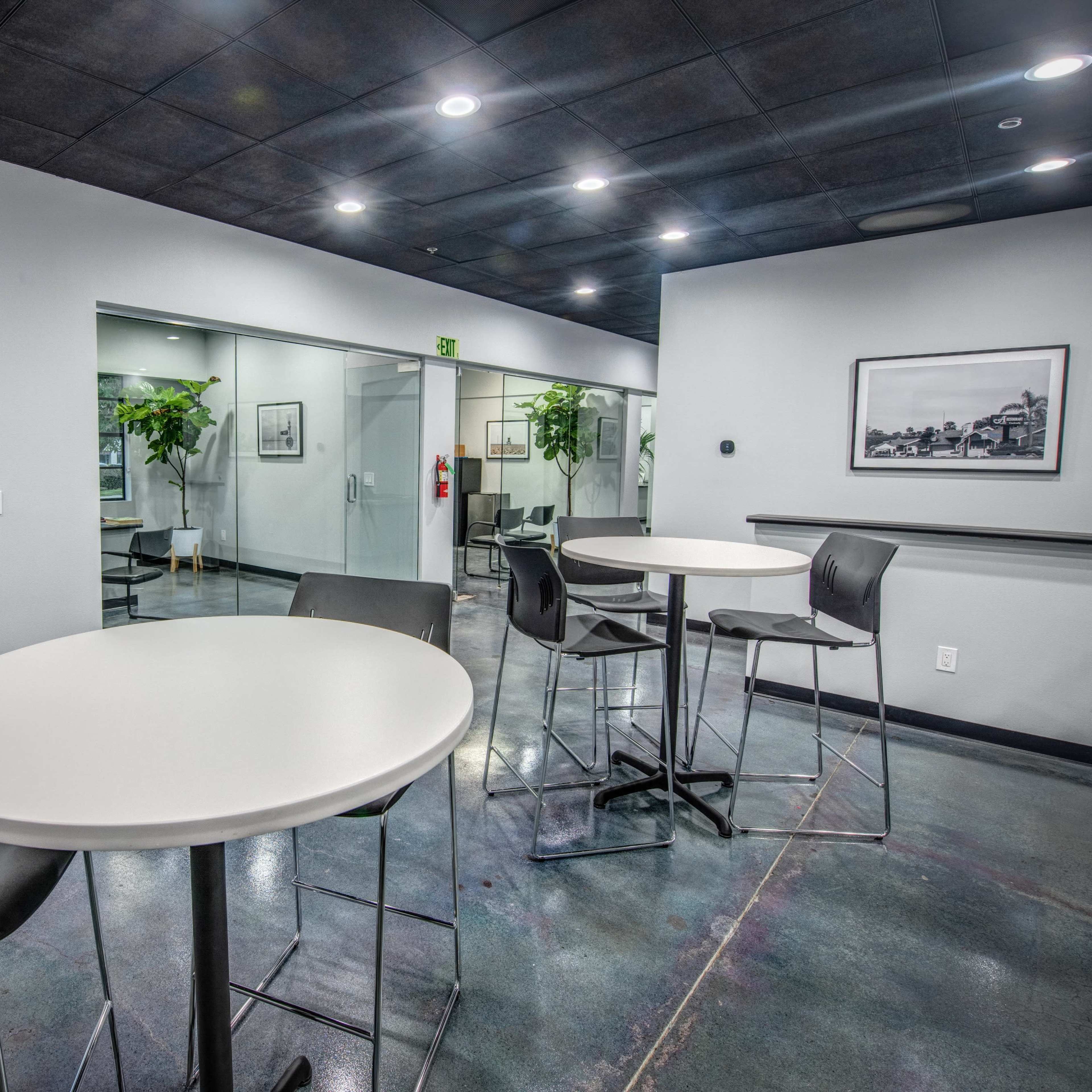 A modern office space features round tables with high stools, surrounded by potted plants and glass-walled meeting rooms.