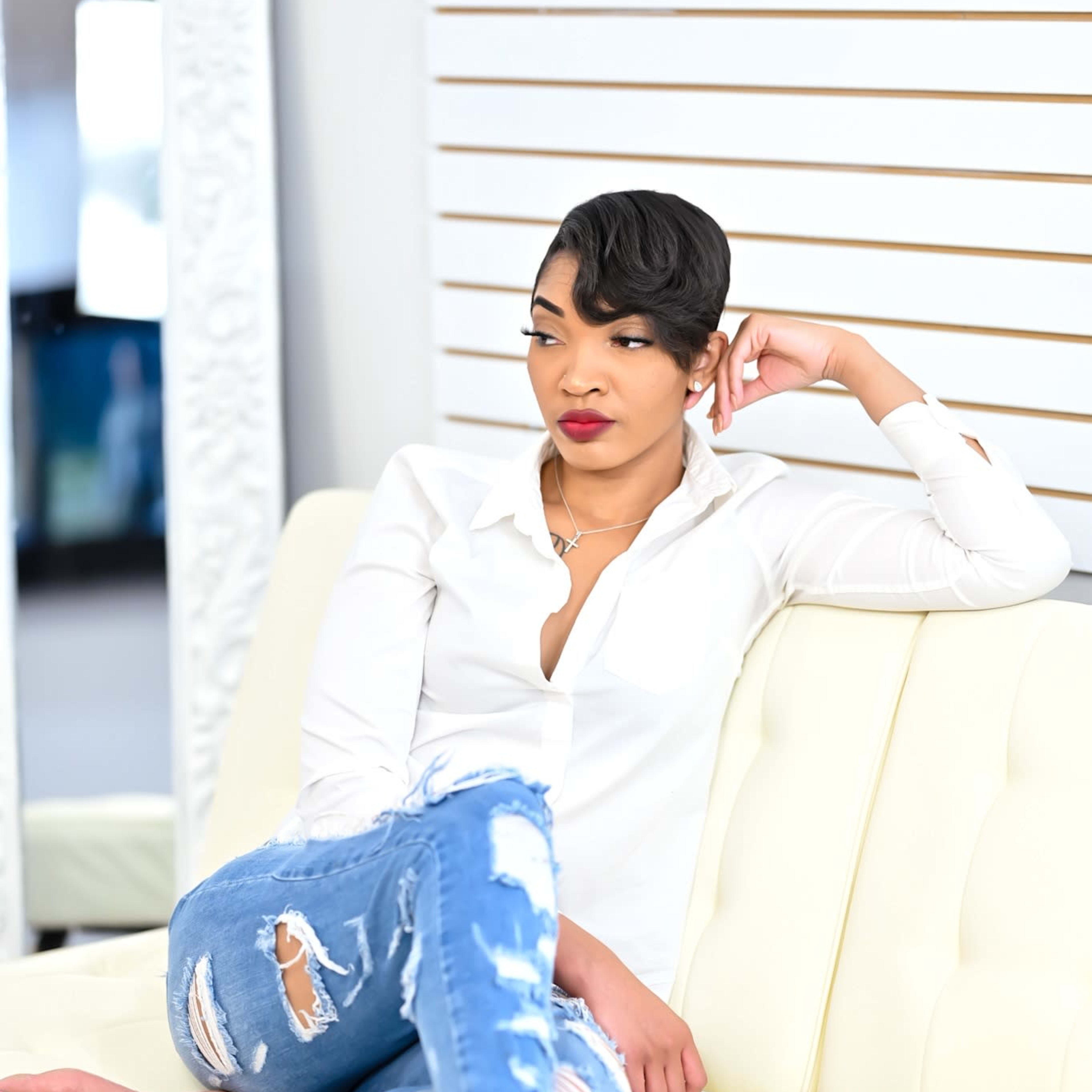 A woman in a white shirt and ripped jeans sits barefoot on a white sofa, leaning against a wall with vertical stripes.