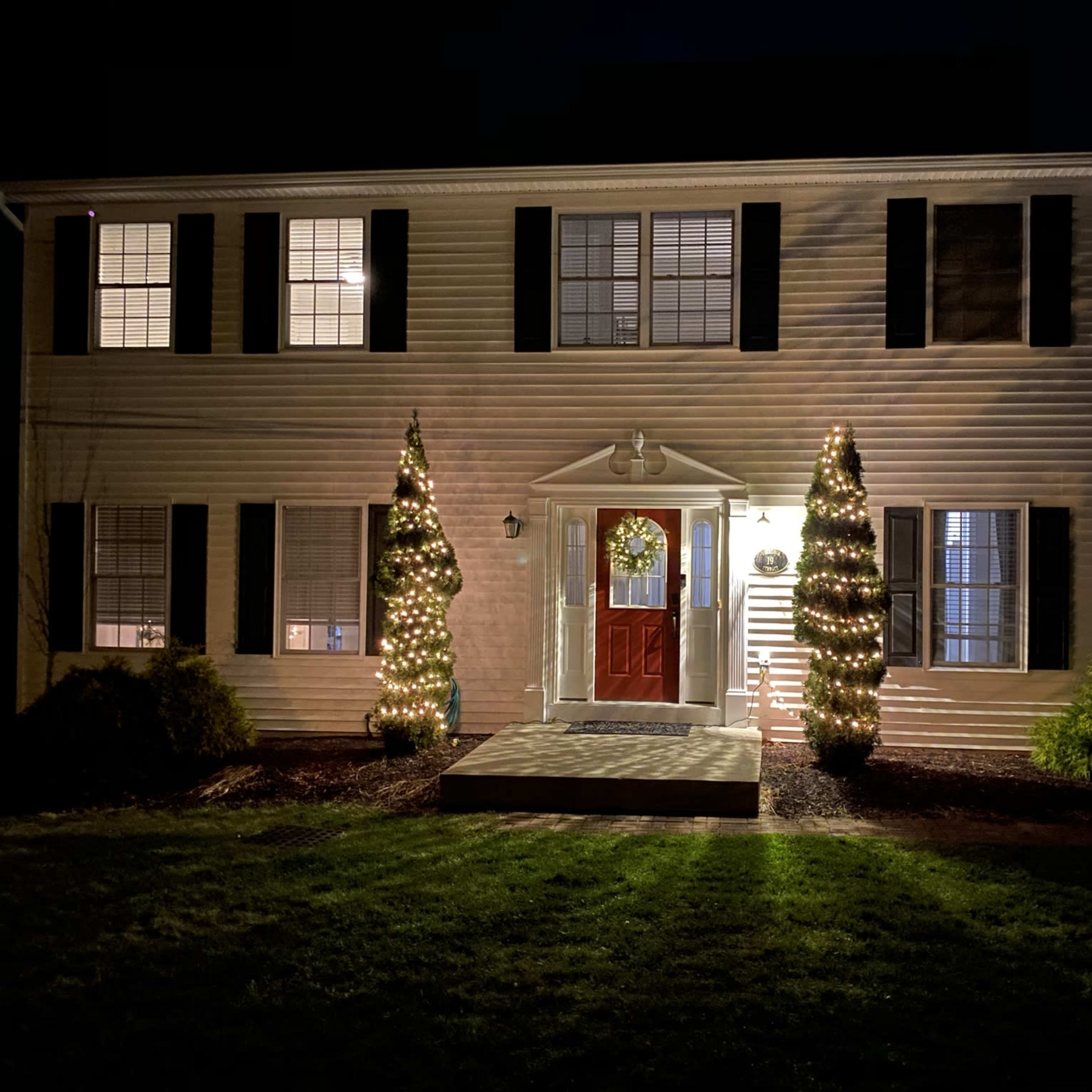 A two-story house is decorated with holiday lights on either side of the front entrance, featuring a wreath on the door and well-lit conifer trees.