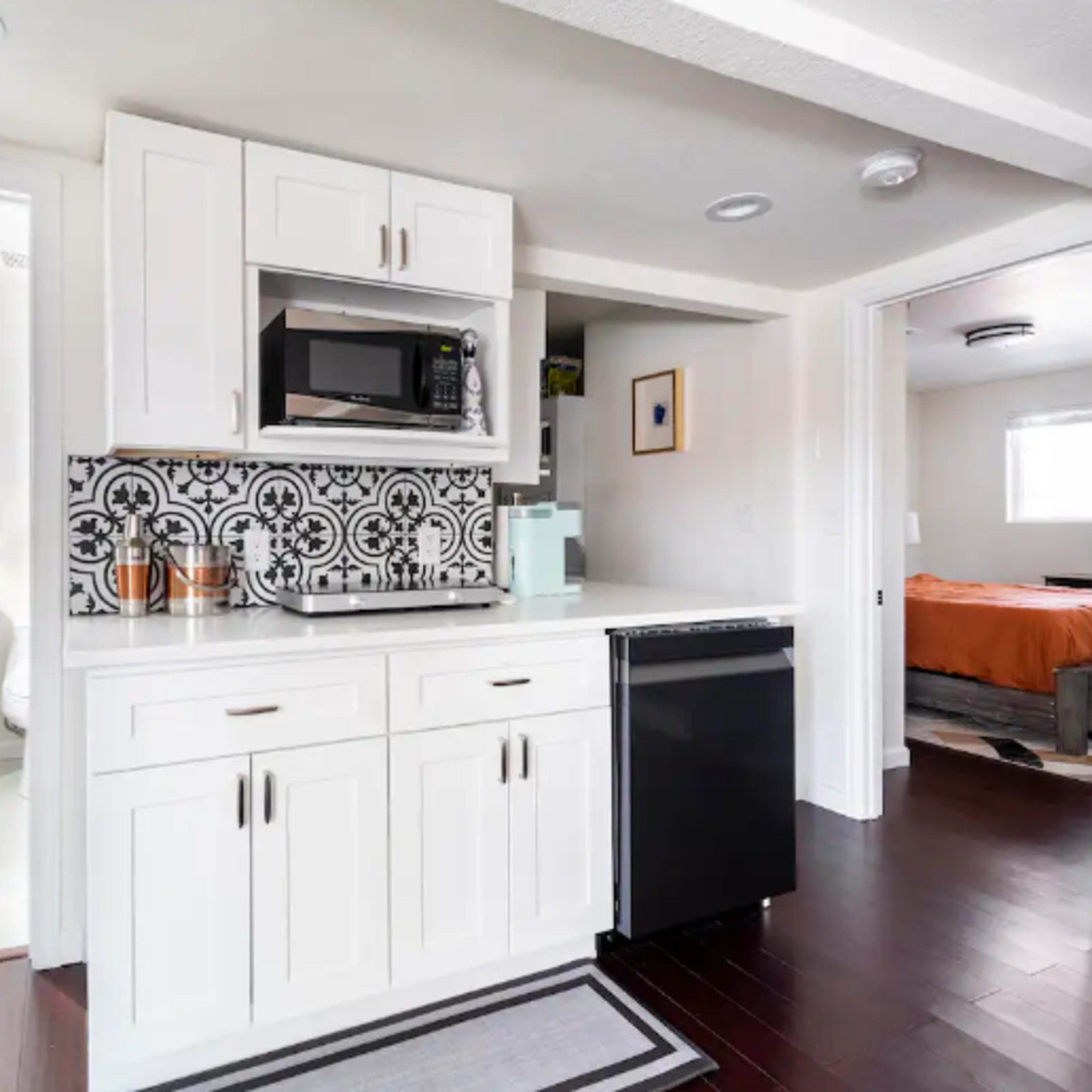 A modern kitchen with white cabinets, a microwave, and a black refrigerator, adjacent to a bathroom with a sink and a doorway leading to a bedroom.