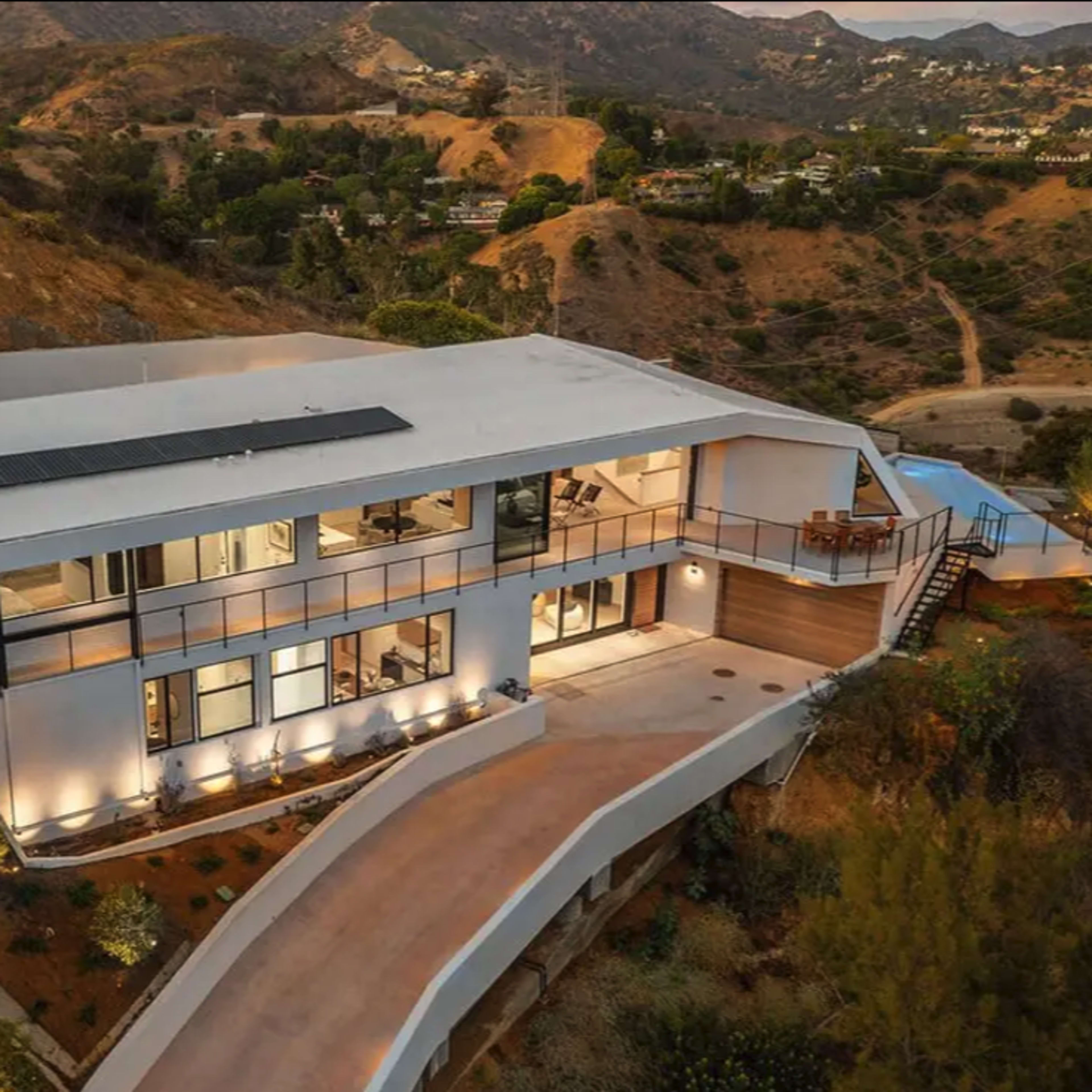 A modern house with large windows and a sloped driveway is situated on a hillside, surrounded by greenery and vistas of rolling hills.