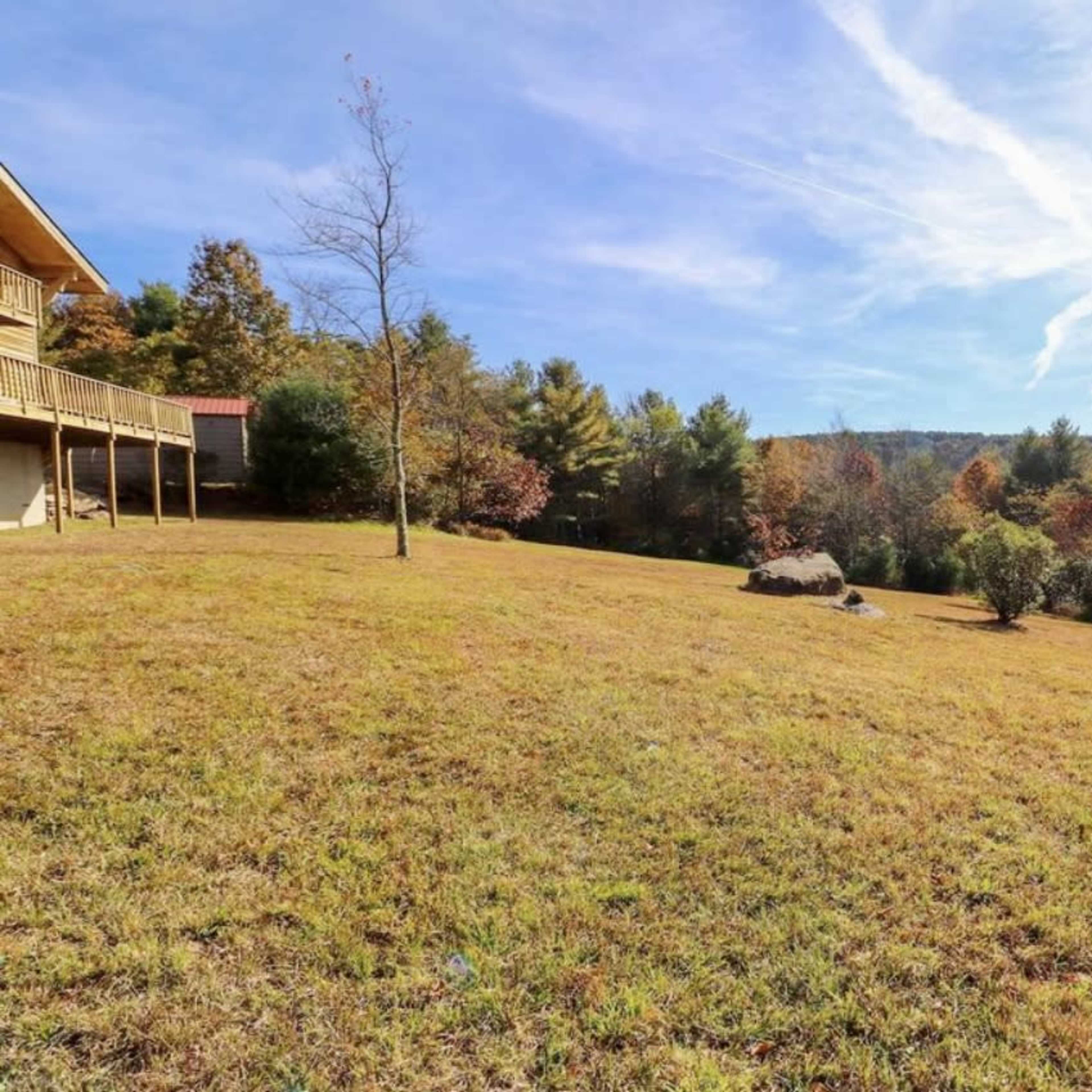 A grassy field slopes gently down towards a wooded area, with a large rock positioned in the center and a house visible on the left.
