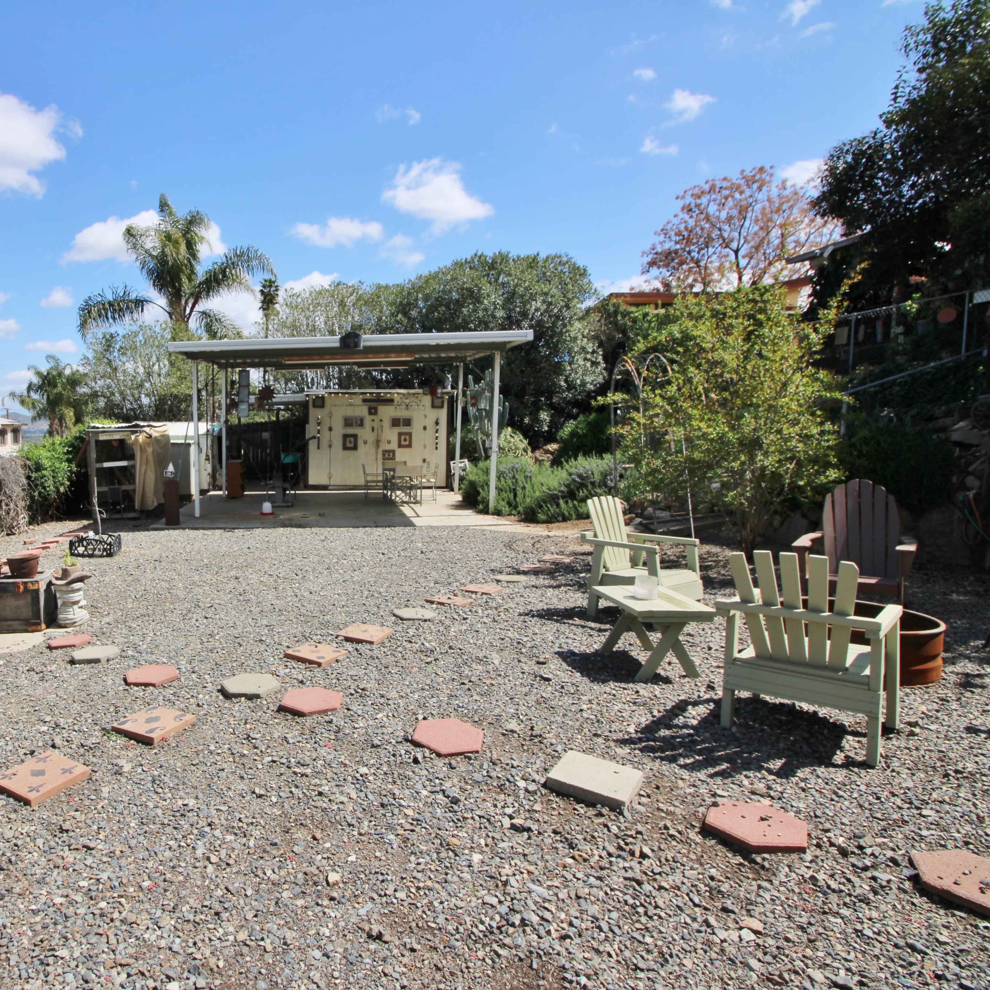 A gravel patio features seating areas with white and green chairs, leading to a shaded structure surrounded by plants and trees.