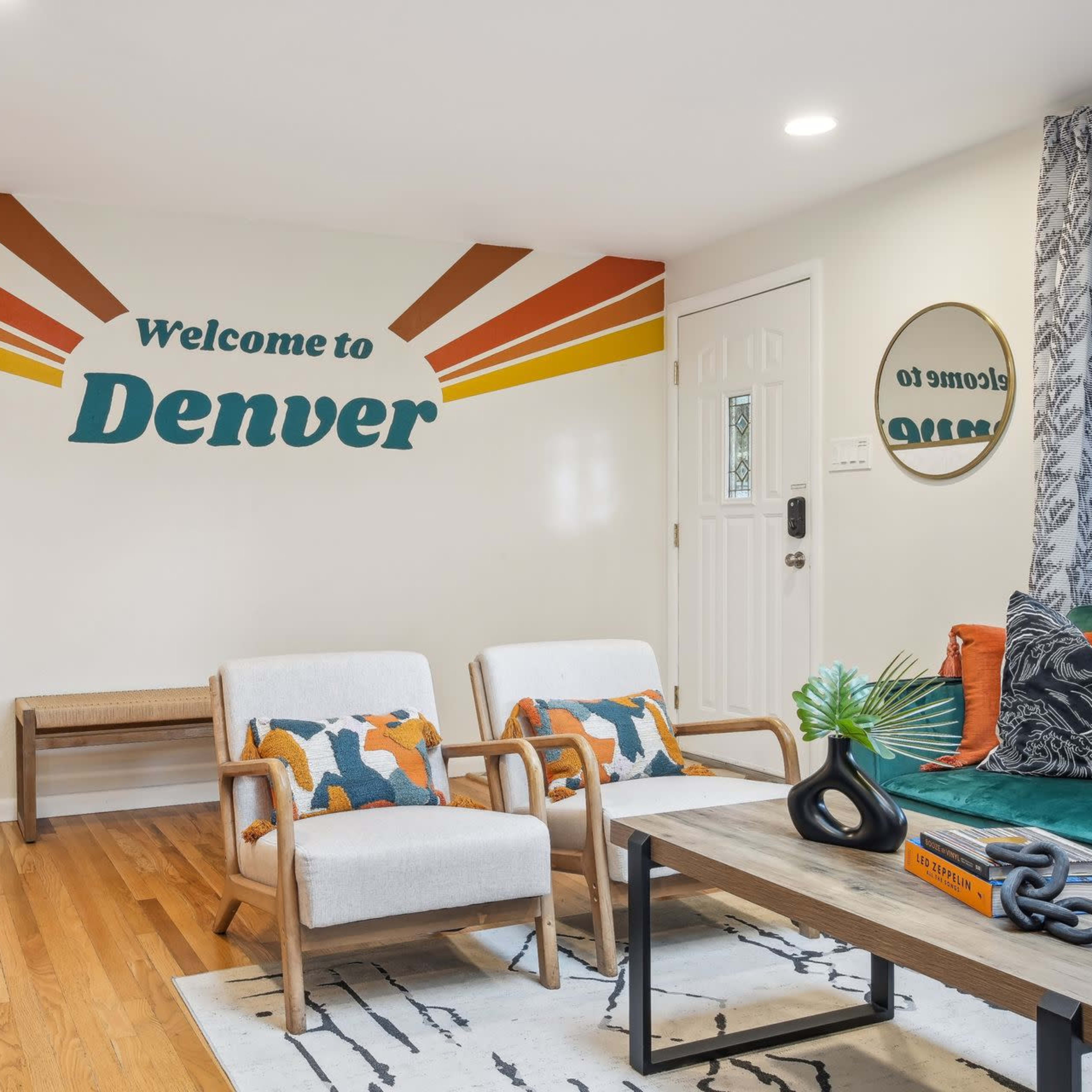 The living room features a welcoming mural that reads "Welcome to Denver," paired with modern furniture and natural wooden accents.
