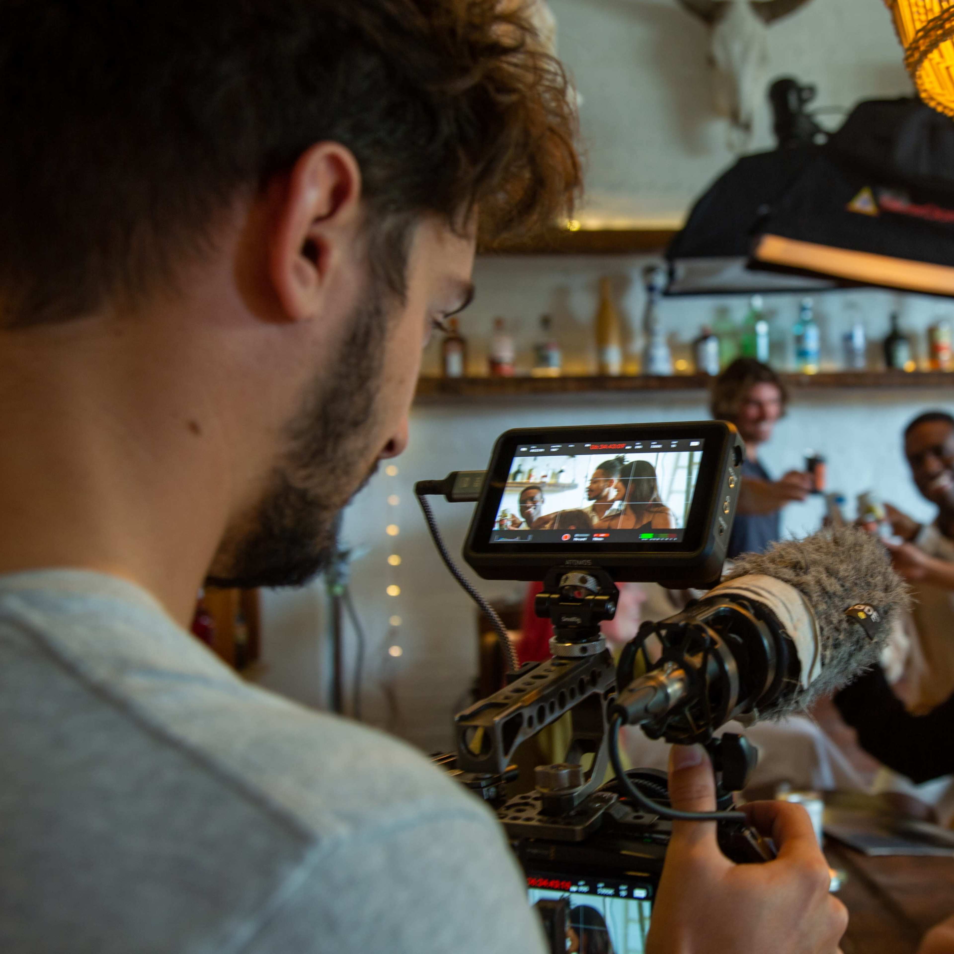 A person operates a camera while filming a group of people gathered around a table in a well-lit room adorned with decorative items.