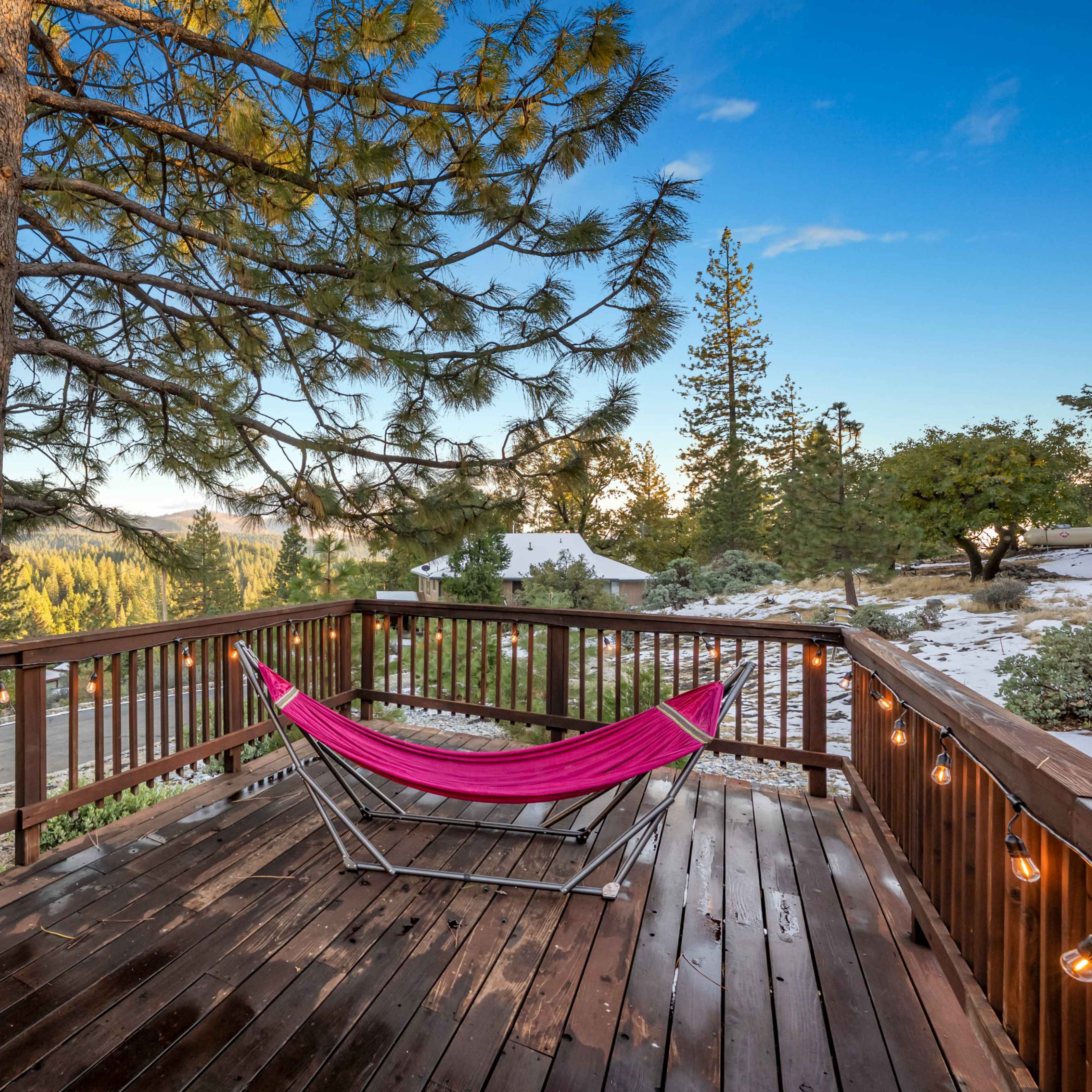 A wooden deck with a pink hammock overlooks a forested landscape of pine trees and distant mountains.