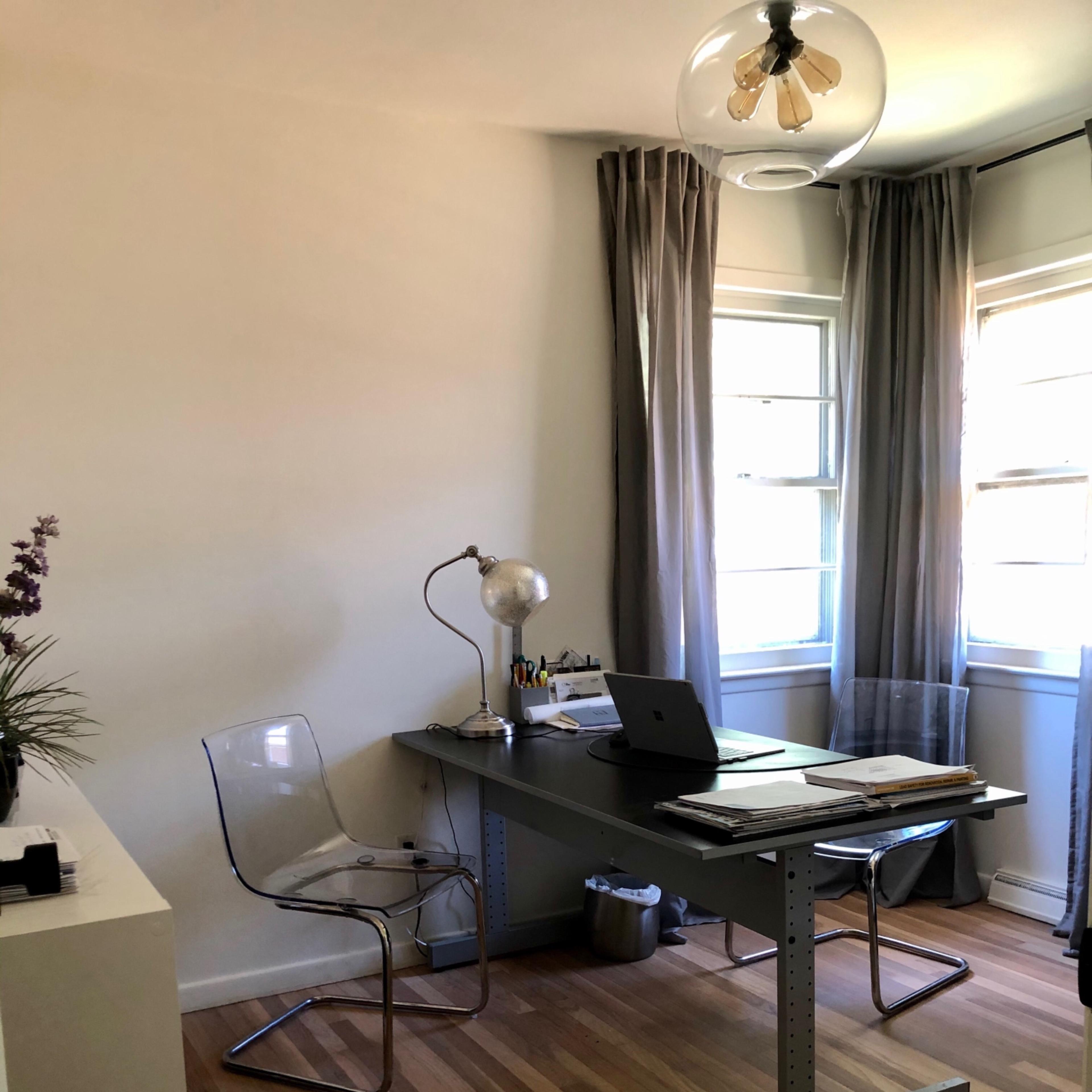 A modern office space features a clear acrylic chair, a sleek desk with a laptop, and natural light filtering through sheer curtains.