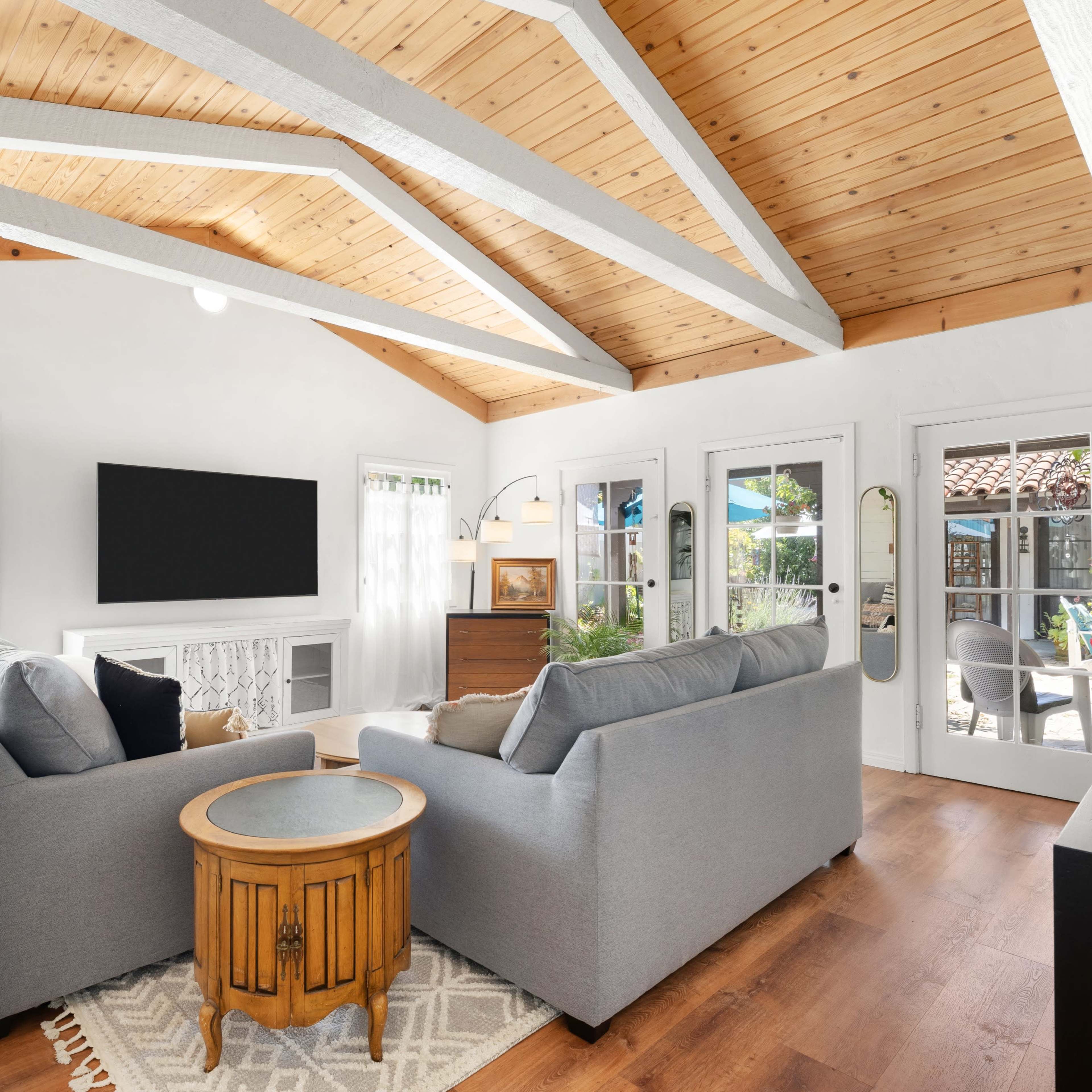 A cozy living room features two gray sofas, a round wooden table, and a large TV mounted on a white wall with exposed wooden beams above.