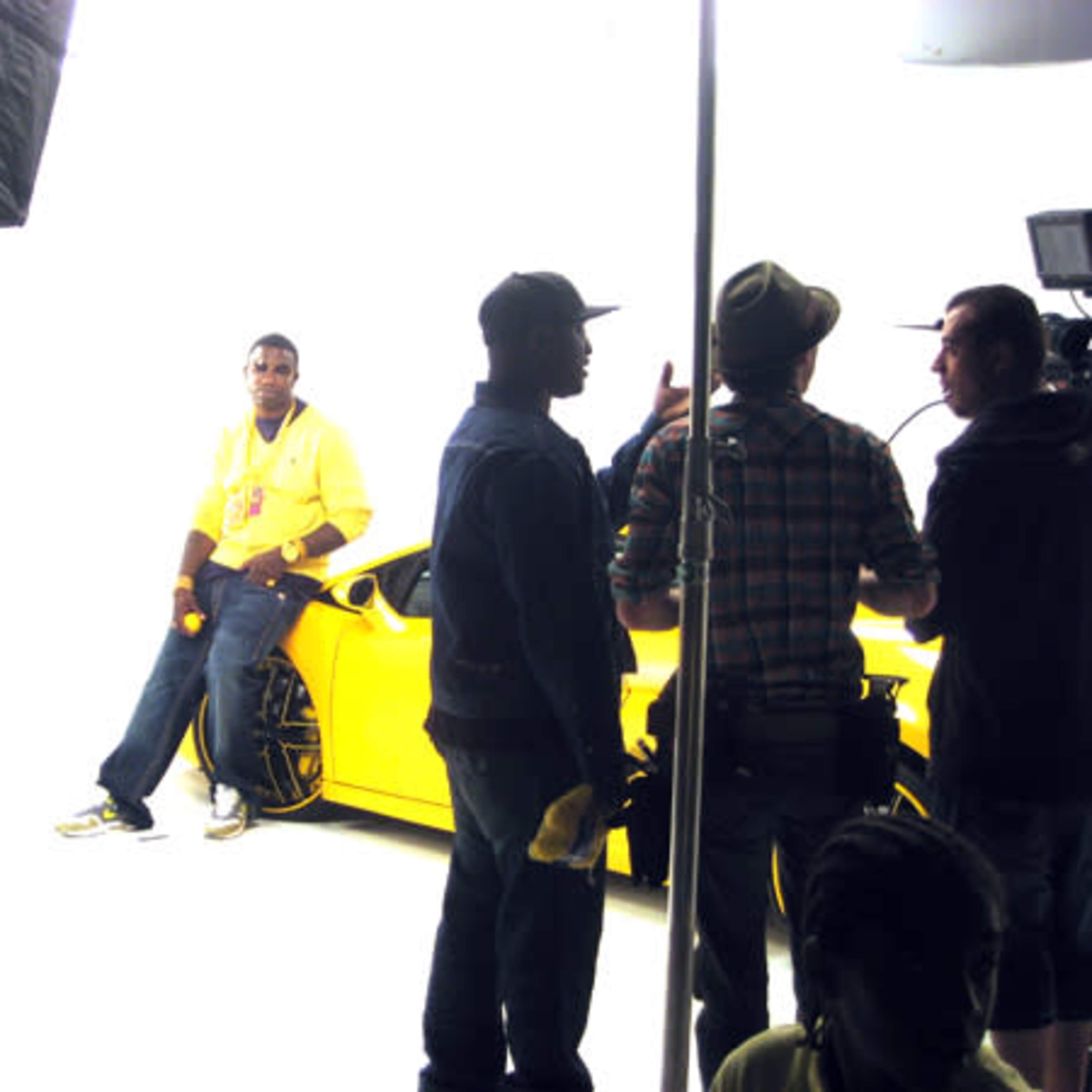 A yellow sports car is featured in a photo shoot with a model posing nearby while a group of crew members discuss in the background.