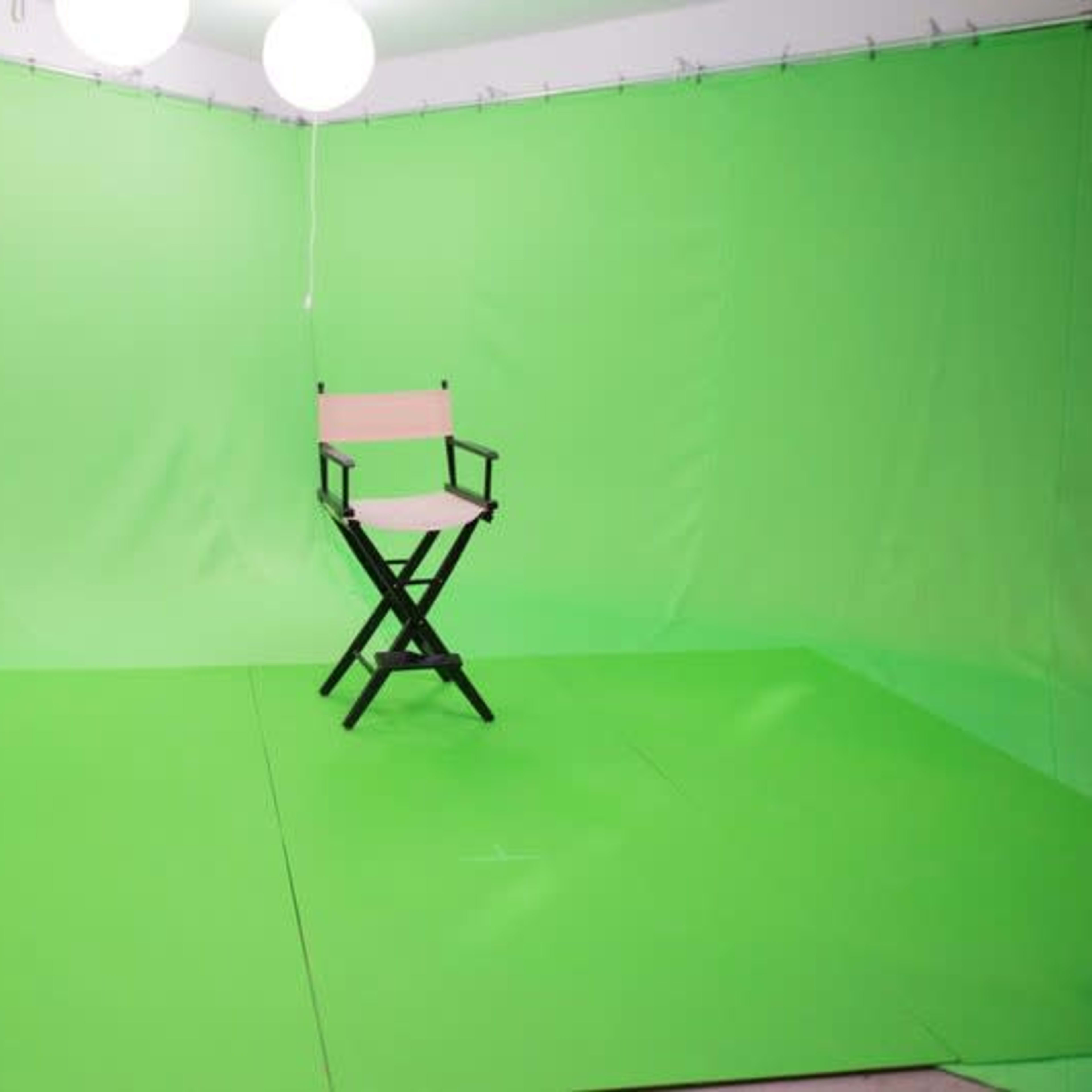 An empty green screen studio with a director's chair positioned on a green floor, illuminated by hanging lights.