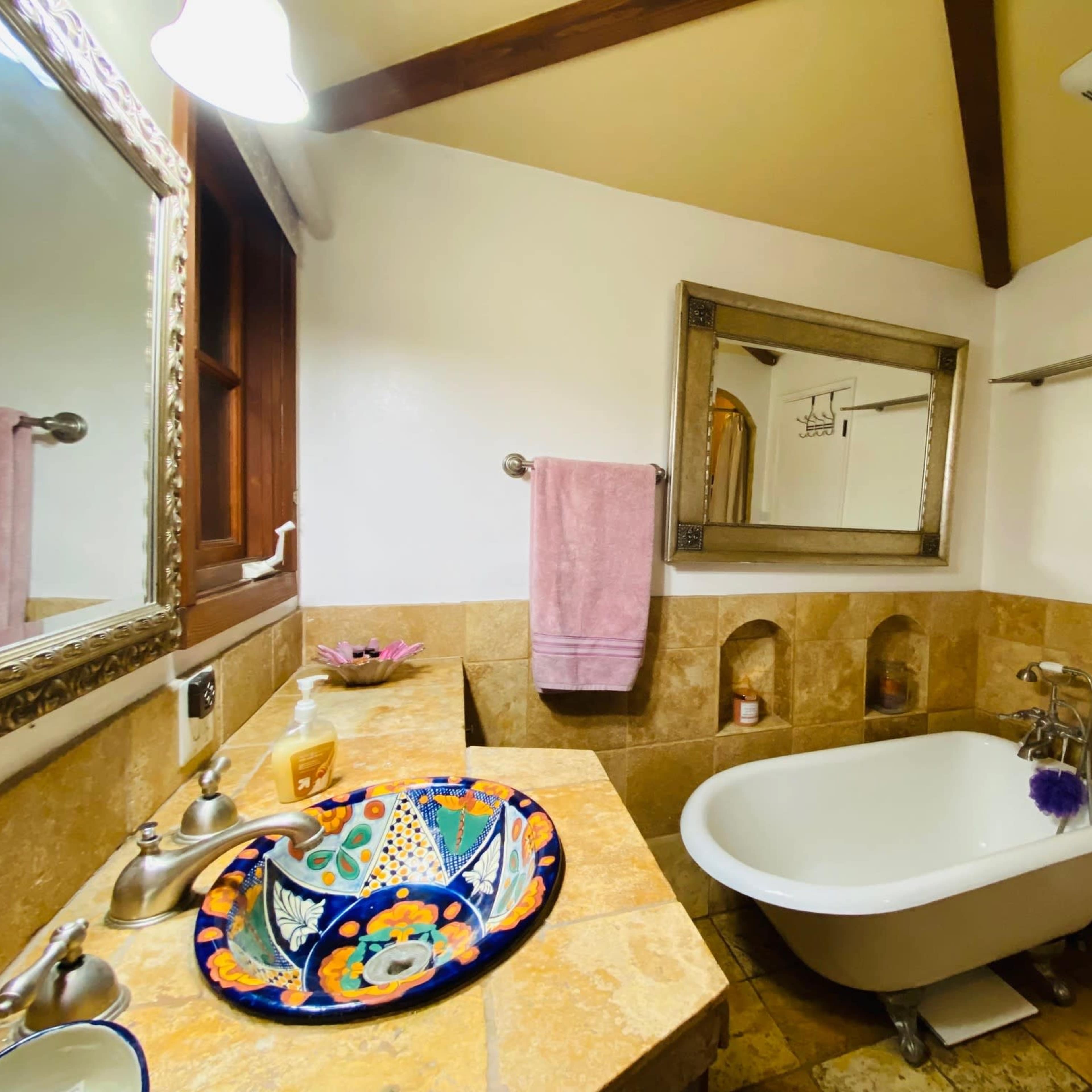 A bathroom with a vintage-style freestanding bathtub, a decorative wash basin, a large mirror, and beige tiled walls.