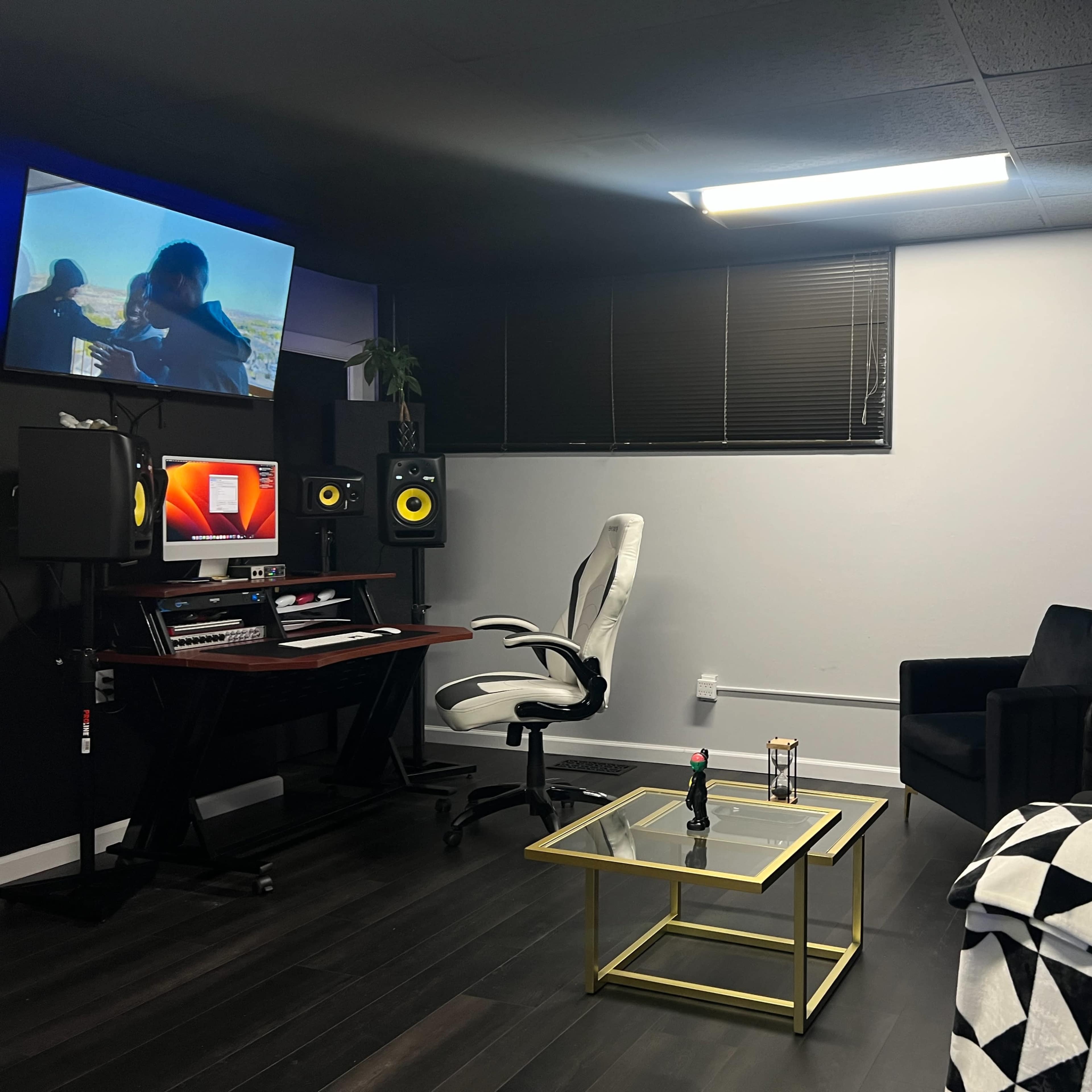 A modern music studio with a desk equipped with audio equipment, a comfortable chair, a couch, a golden coffee table, and a wall-mounted screen displaying visuals.