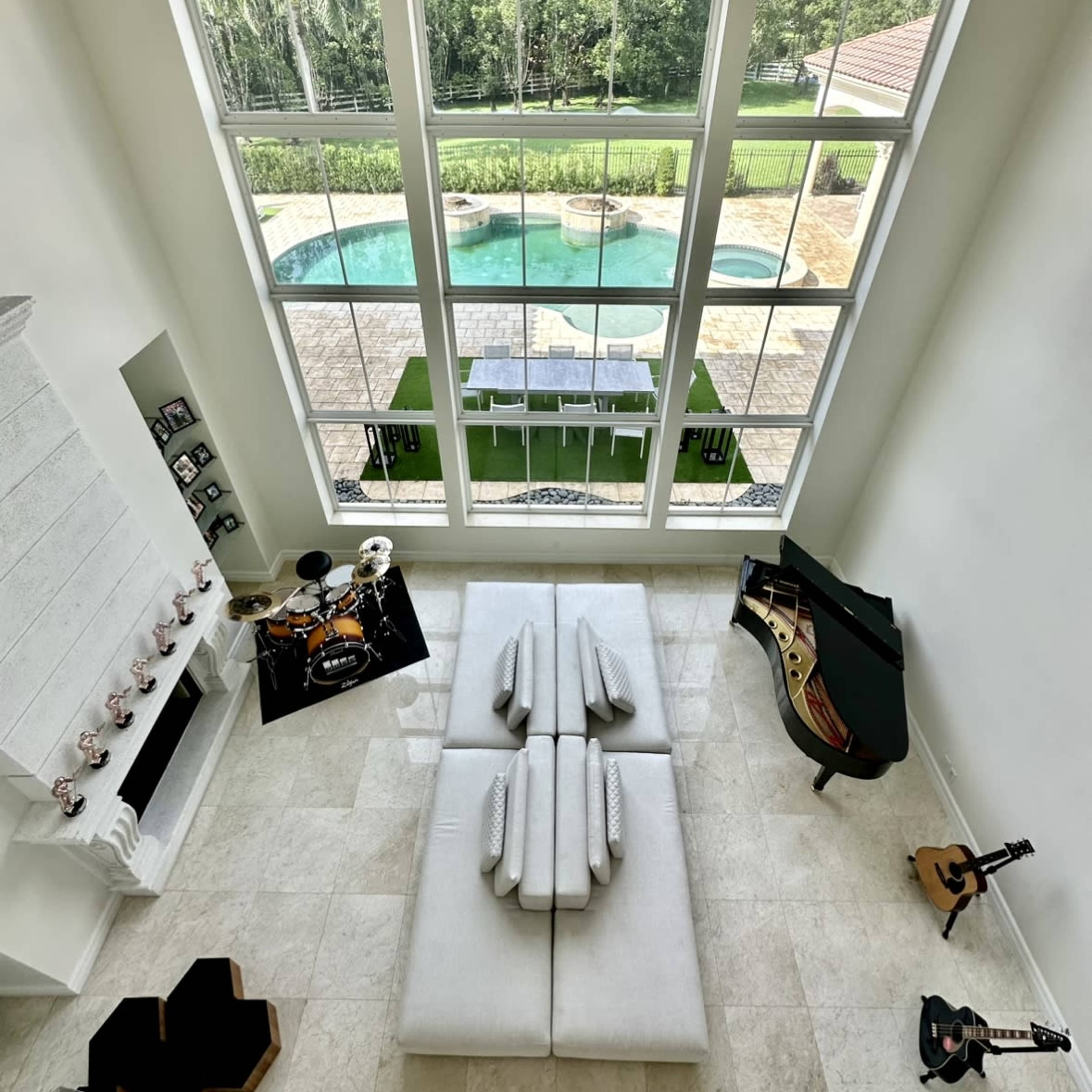 A spacious living room features large windows overlooking a pool and patio area, with a piano, guitar, and drum set arranged indoors.