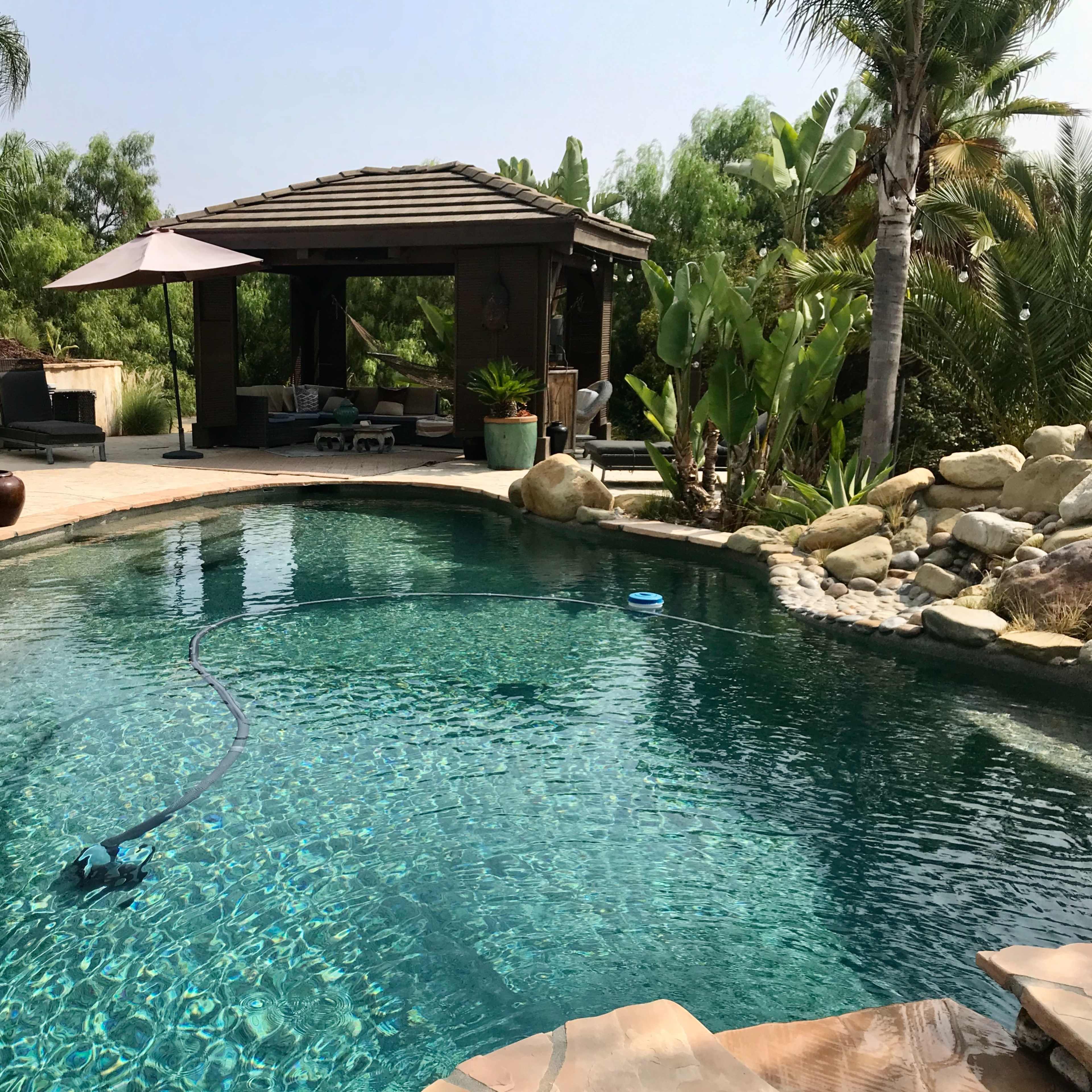 A clear swimming pool surrounded by lush greenery and a shaded gazebo.