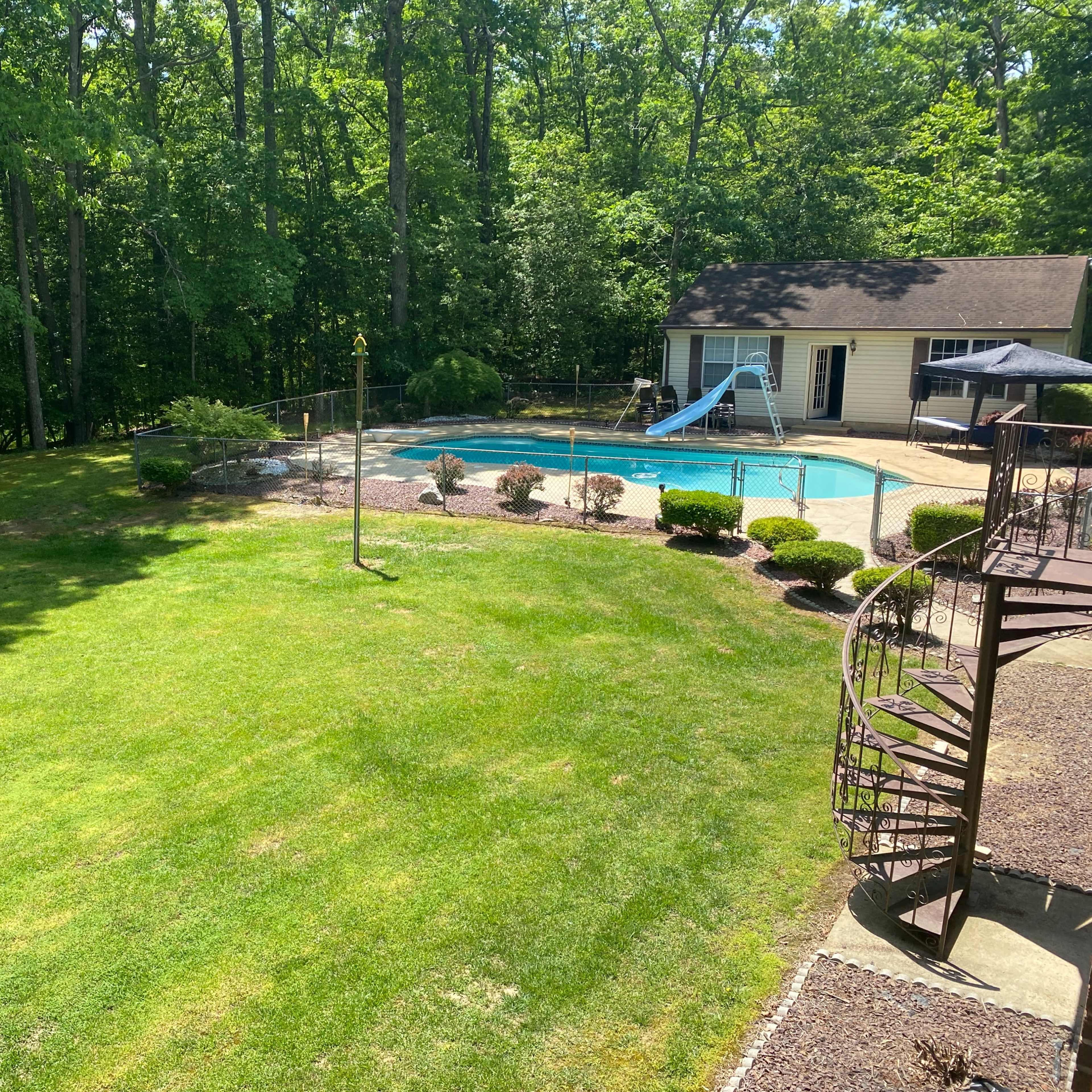 A backyard with a swimming pool, a slide, landscaped bushes, and a spiral staircase leading to a deck, surrounded by trees.