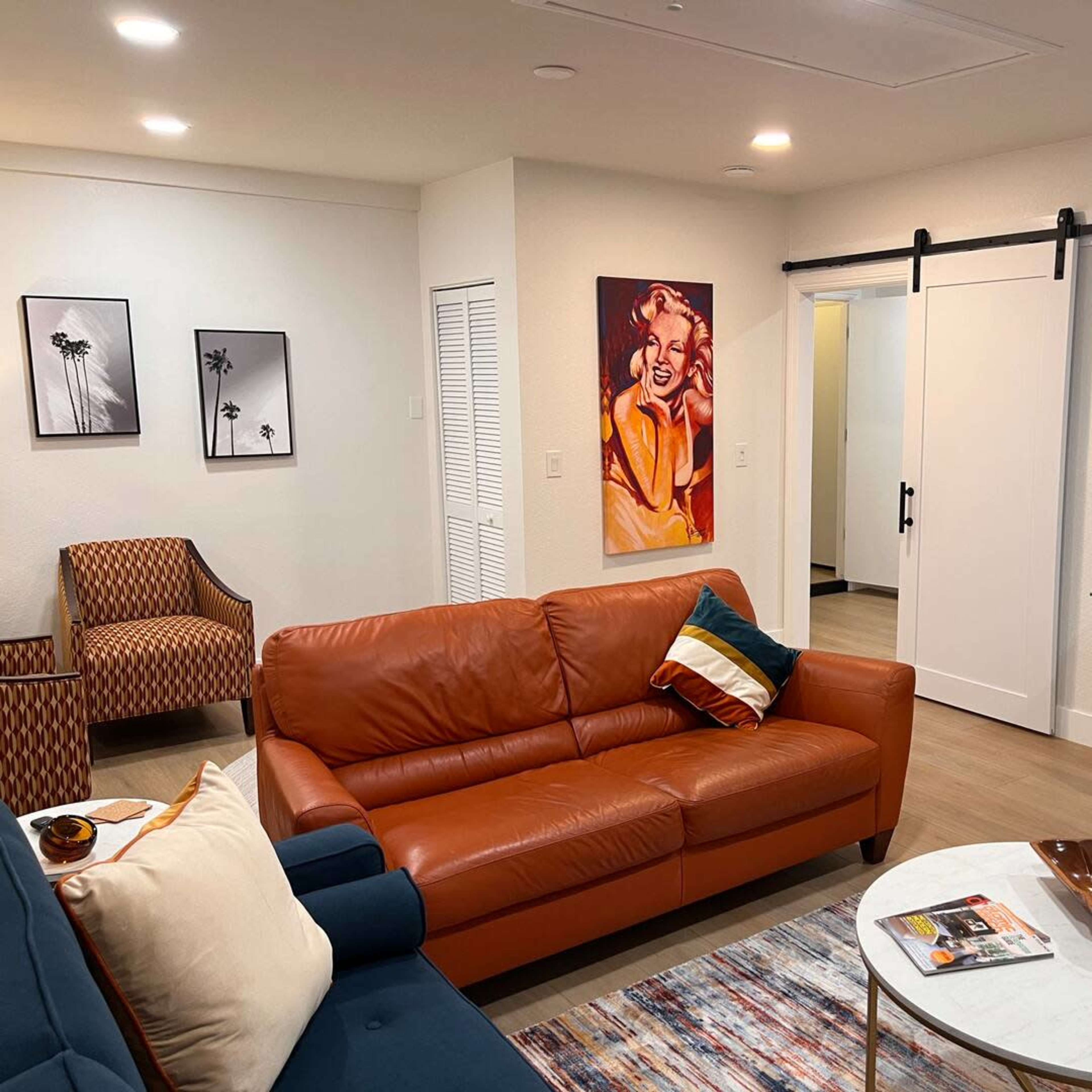 The living room features a brown leather sofa, a blue couch, a coffee table, and wall art, with a sliding door leading to another room.