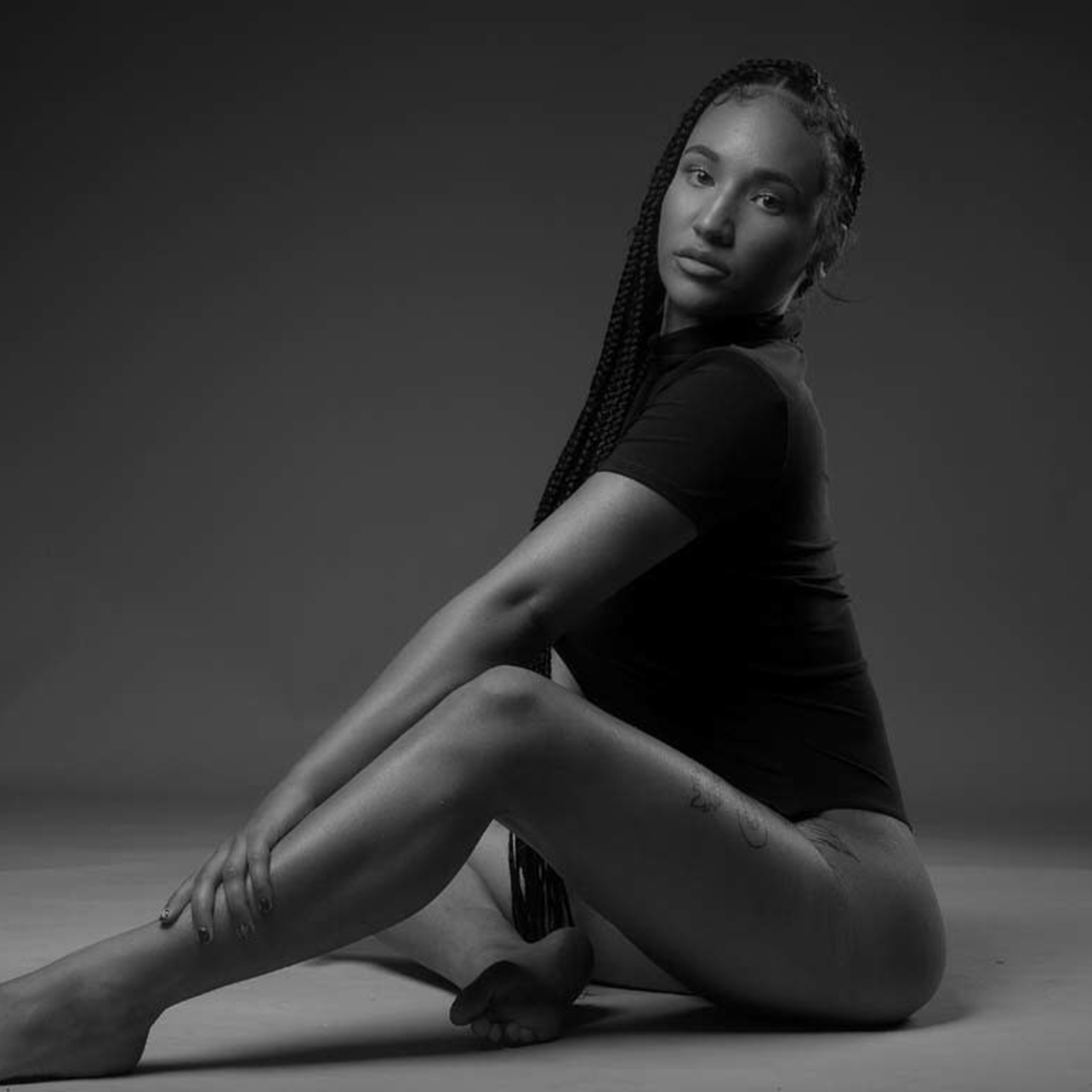 A person with braided hair sits gracefully on the floor, leaning slightly forward with one leg extended and the other bent at the knee.