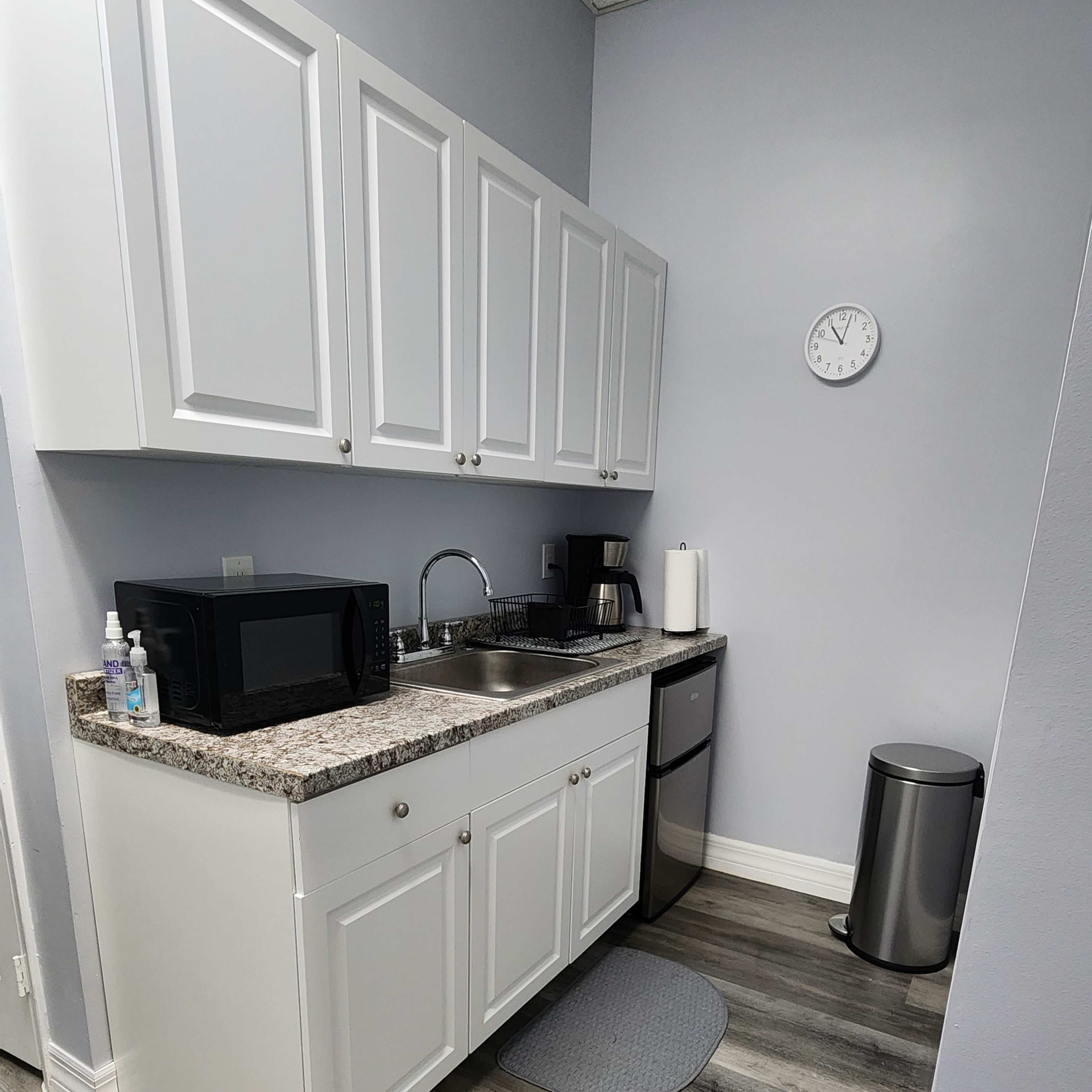 A modern kitchenette with white cabinets, a microwave, a coffee maker, and a stainless steel refrigerator on a gray wall with a clock.