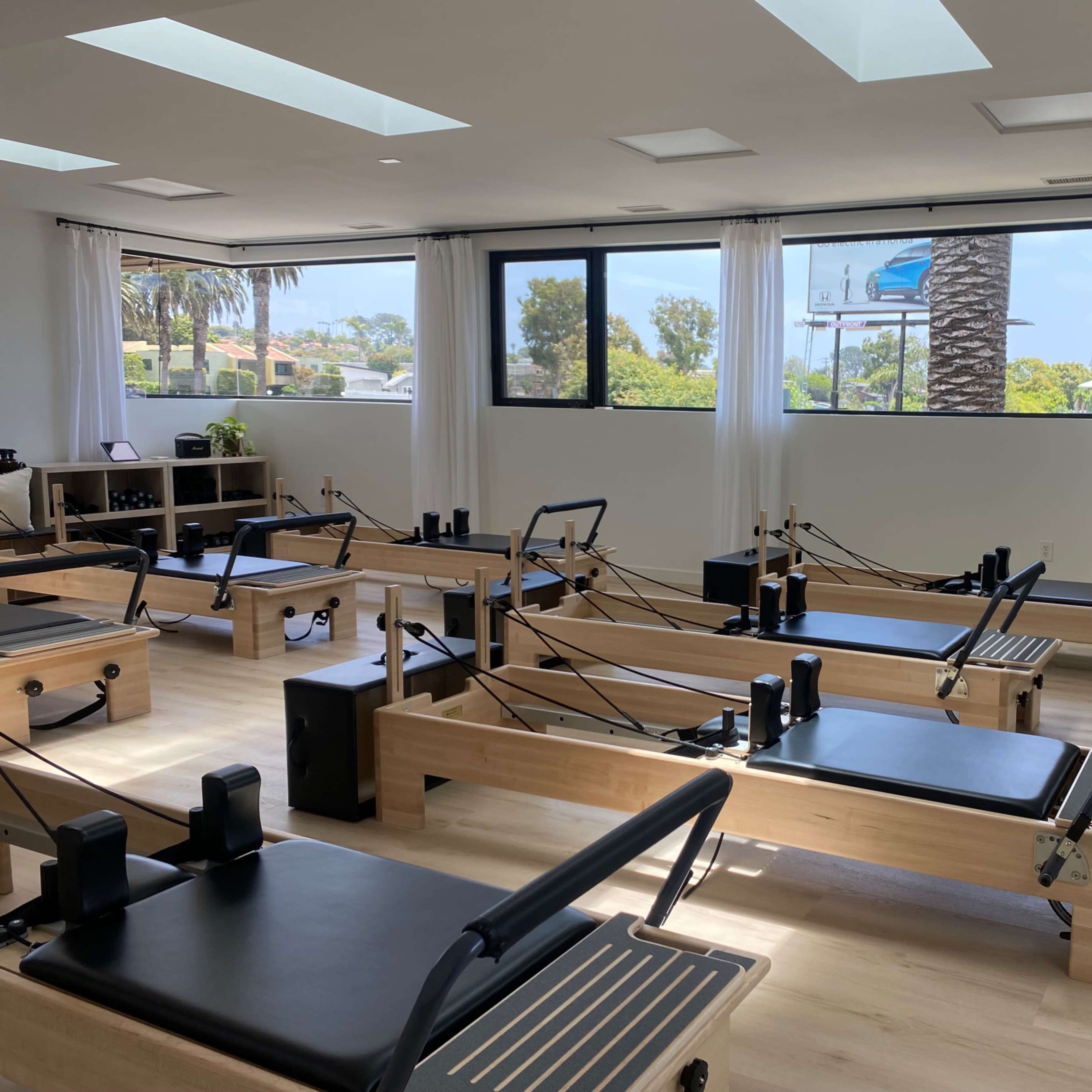 A bright, modern studio equipped with several reformer Pilates machines and large windows overlooking a palm tree-lined view.