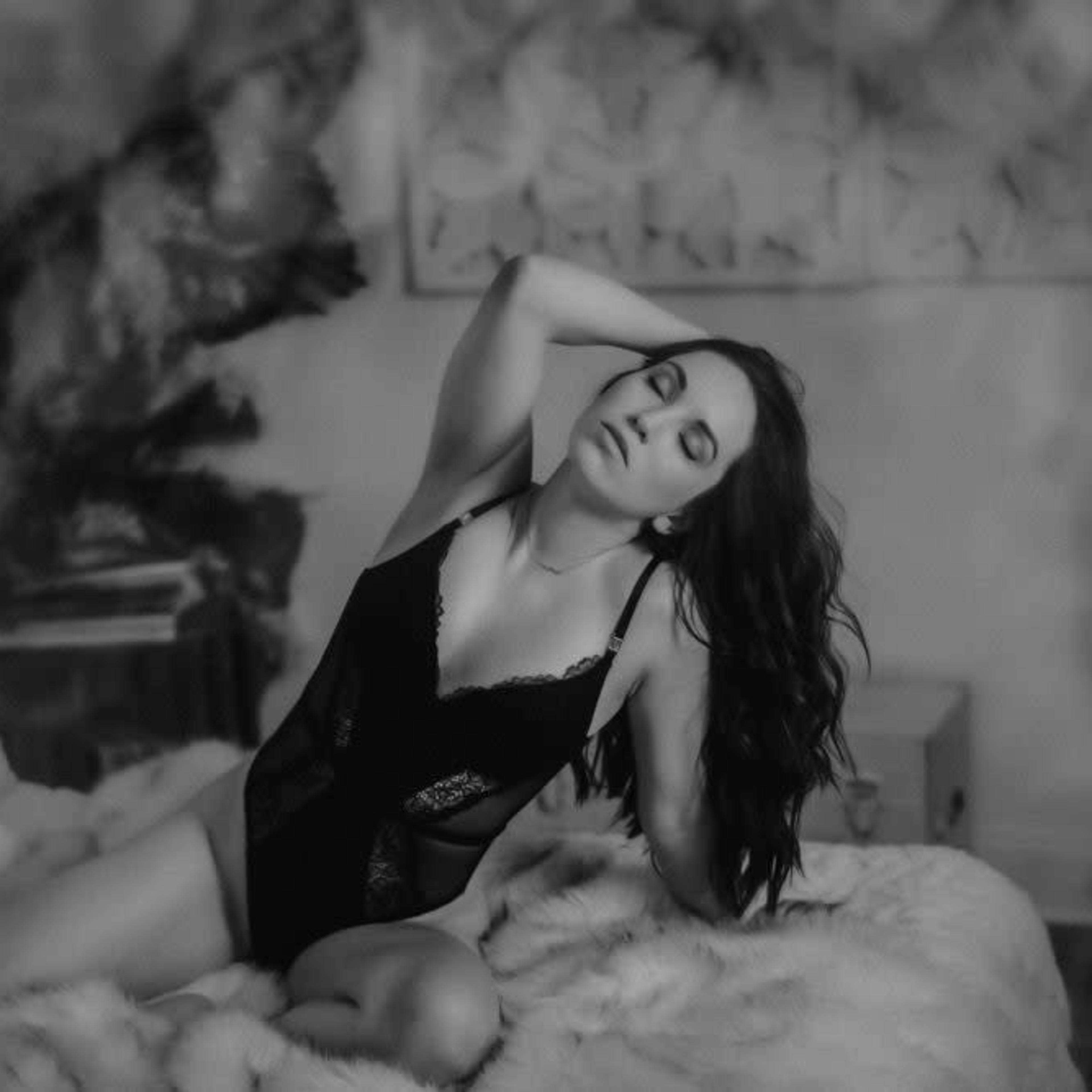 A woman wearing black lingerie poses on a bed with a fluffy blanket in a softly lit room.